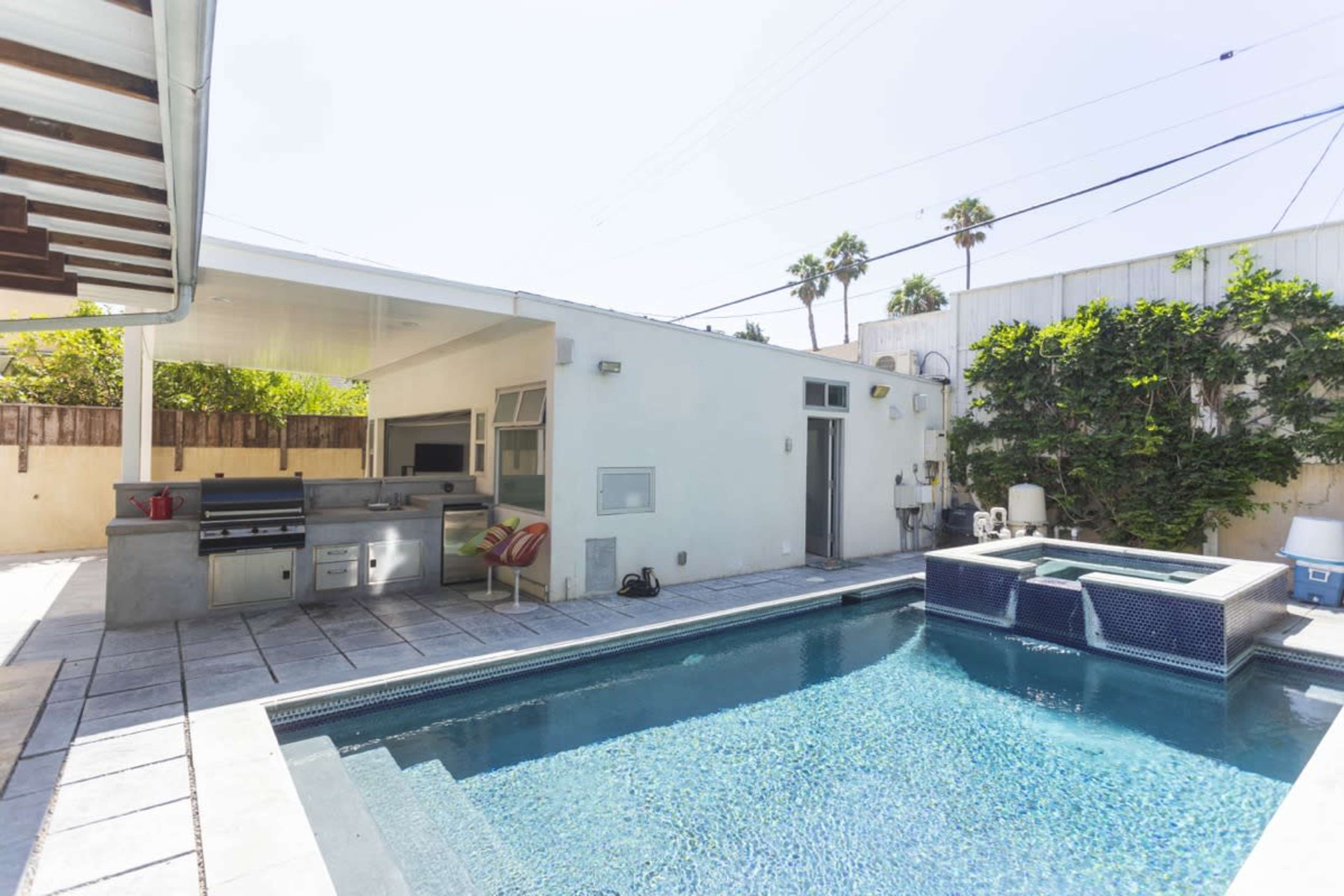 A modern outdoor patio area with a swimming pool, an outdoor kitchen, and surrounding palm trees.
