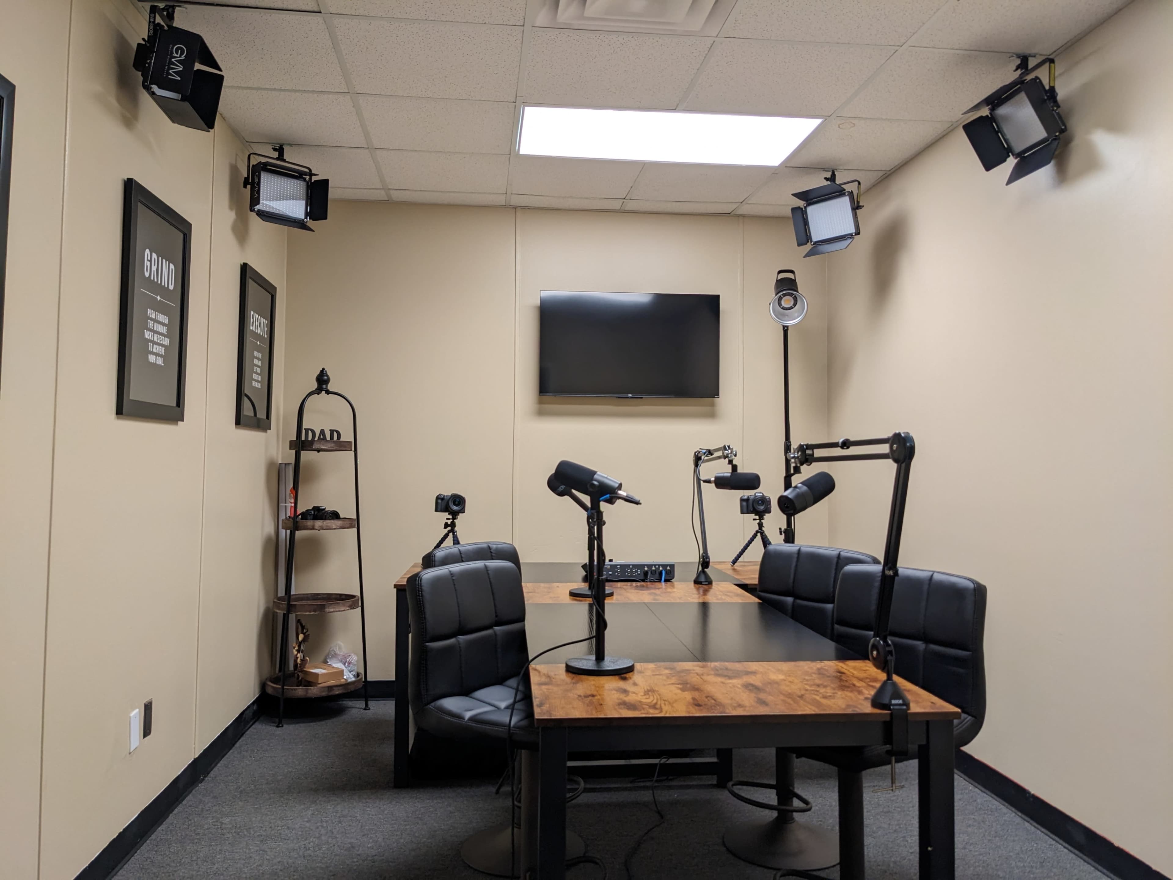 A podcast recording studio features a table with four black chairs, multiple microphones, and a wall-mounted television.