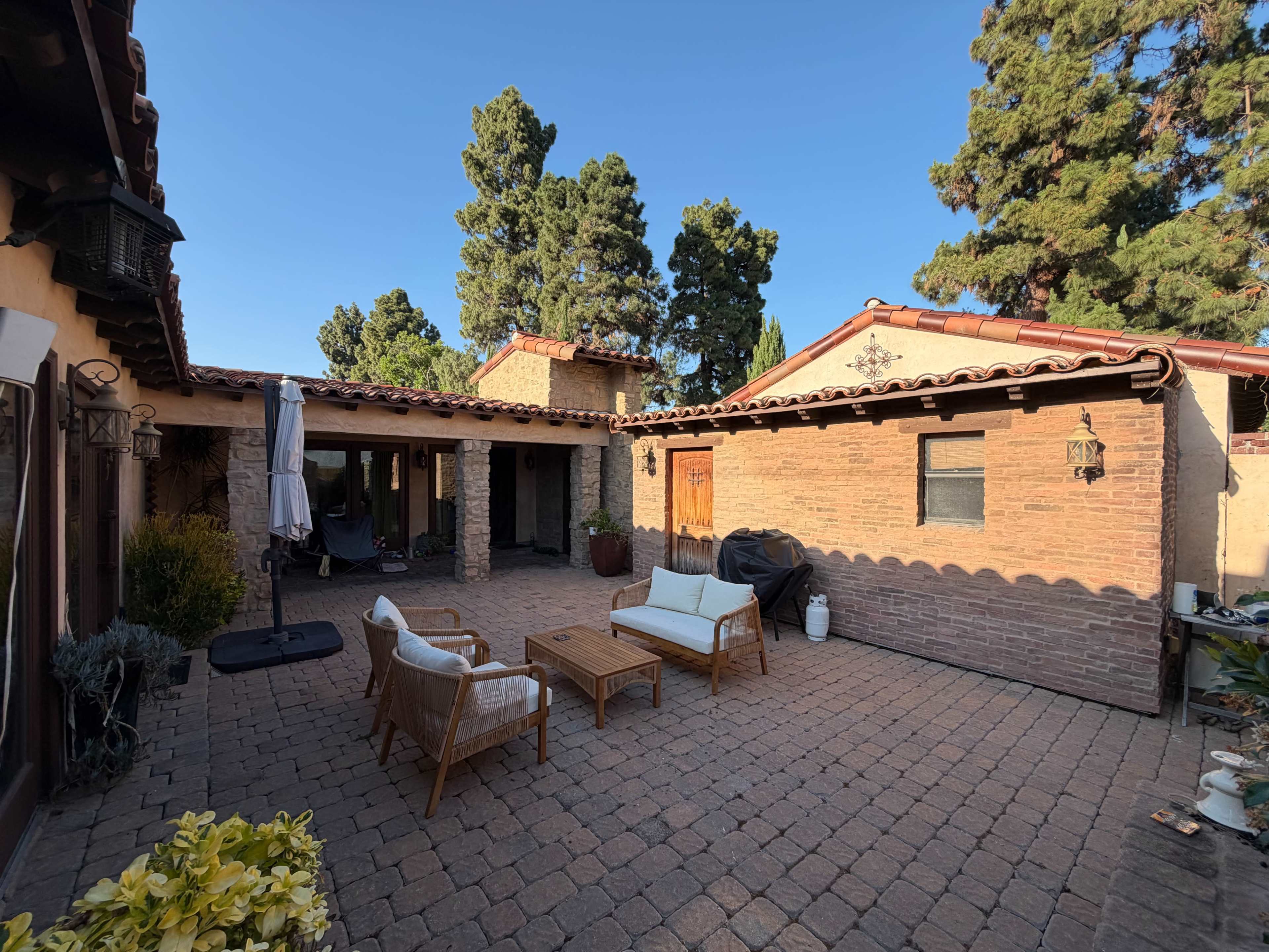 The image shows a spacious outdoor patio surrounded by brick walls and adorned with potted plants, featuring a seating area with a white sofa and wooden coffee table.