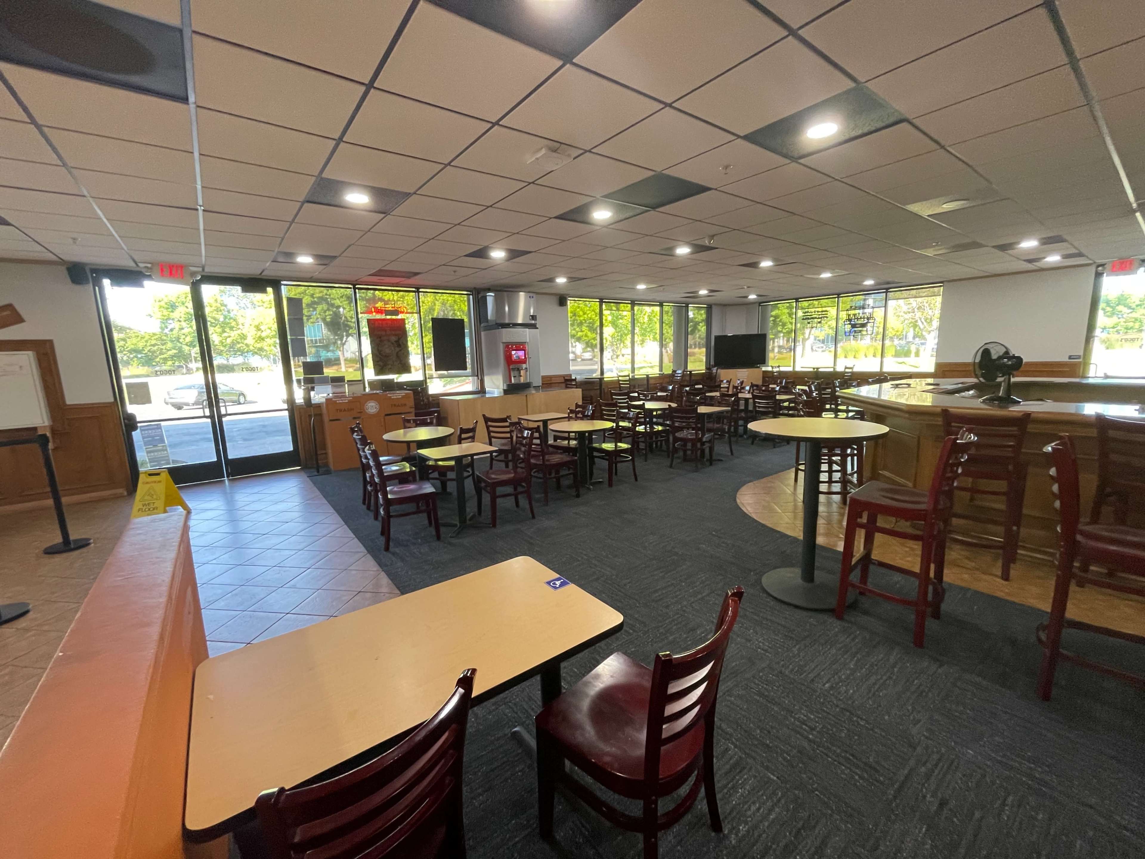 The interior of a dining area with multiple tables and chairs arranged in a spacious layout, large windows allowing natural light, and a service counter in the background.