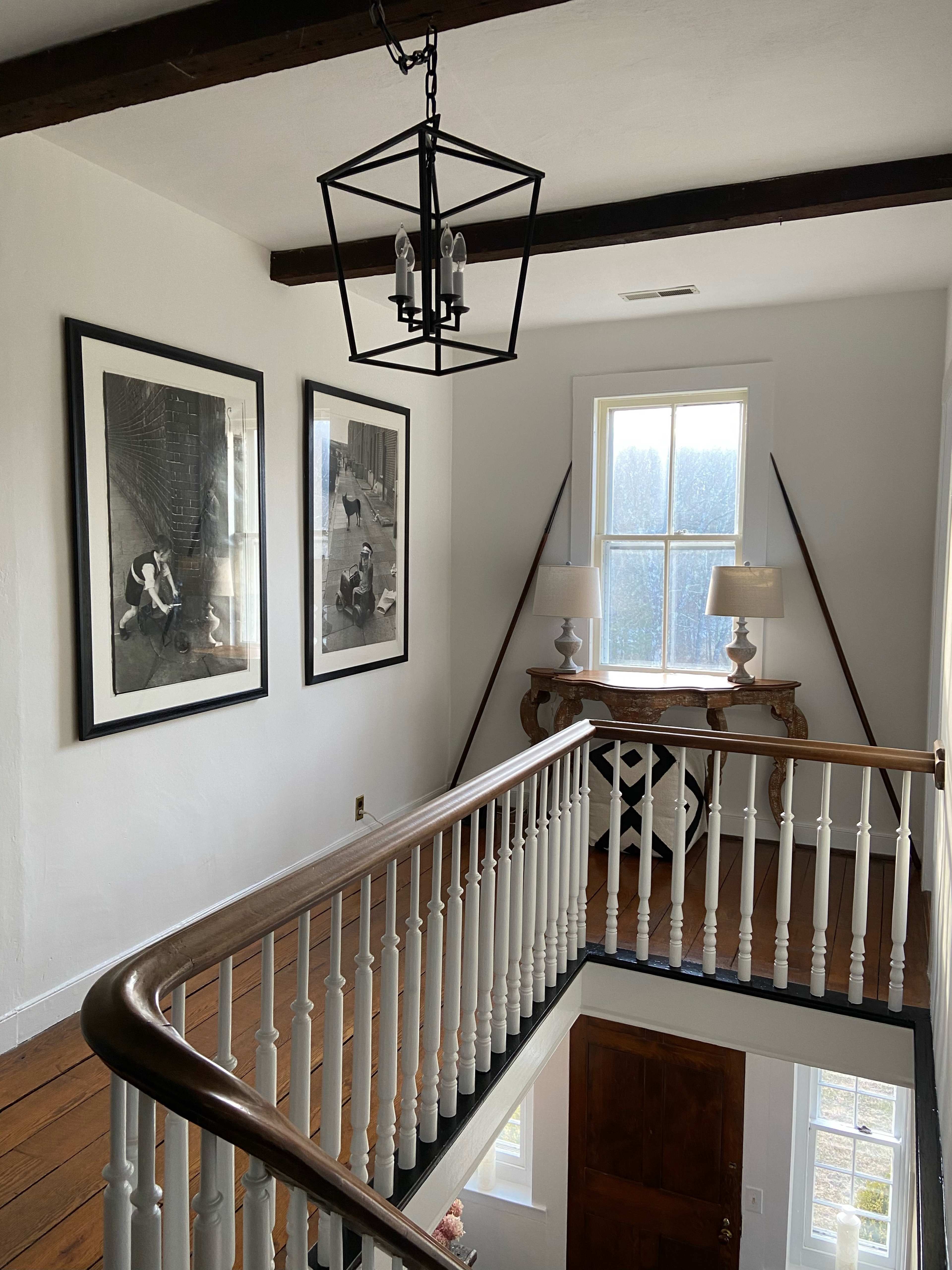 A well-lit upstairs landing with two large framed artworks on the wall, a console table with lamps, and a view of a door and staircase railing.