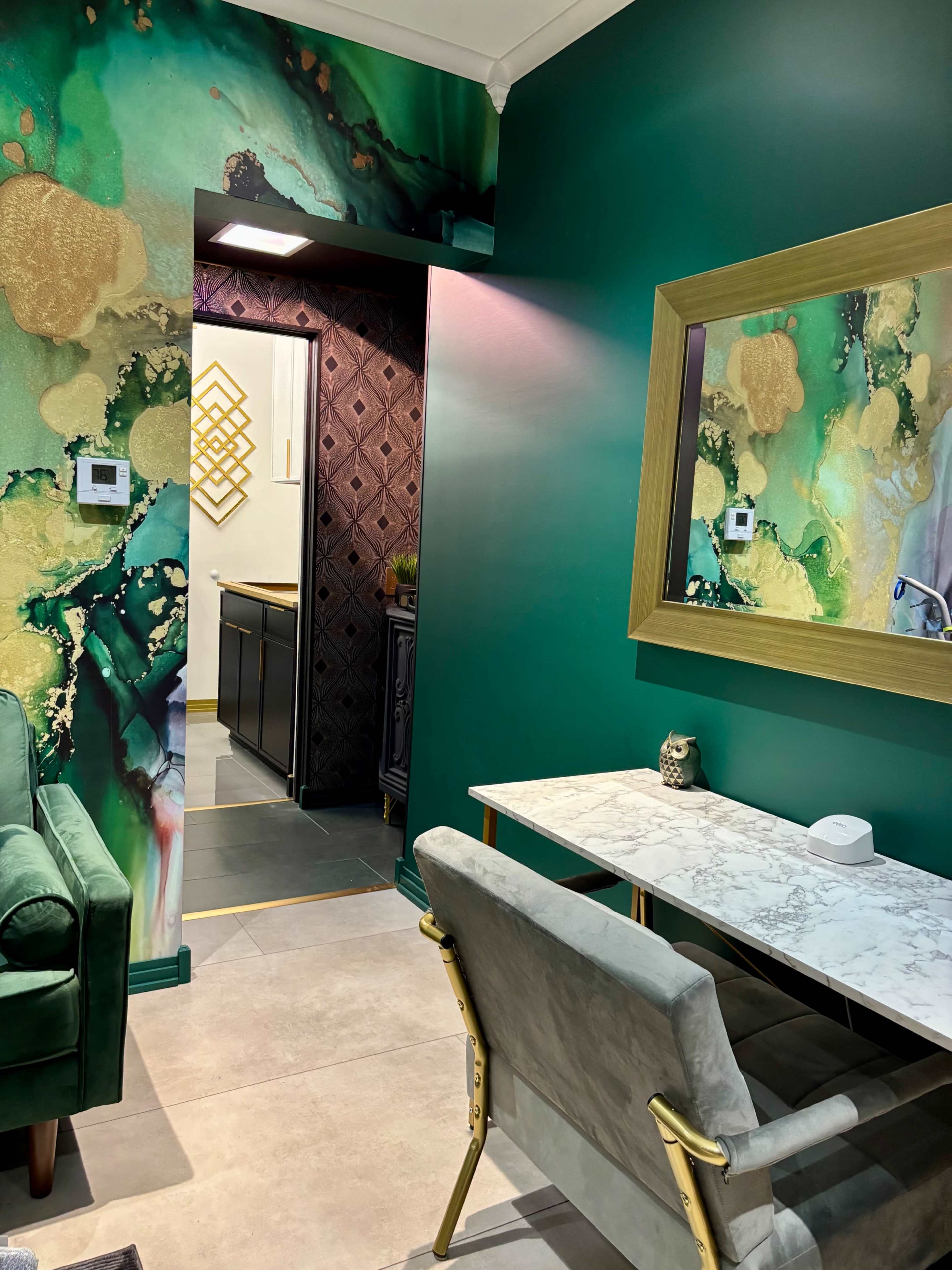 The image shows a stylish interior featuring a green wall with abstract art, a marble-top desk, a gold-framed mirror, and a doorway leading to a kitchen area.