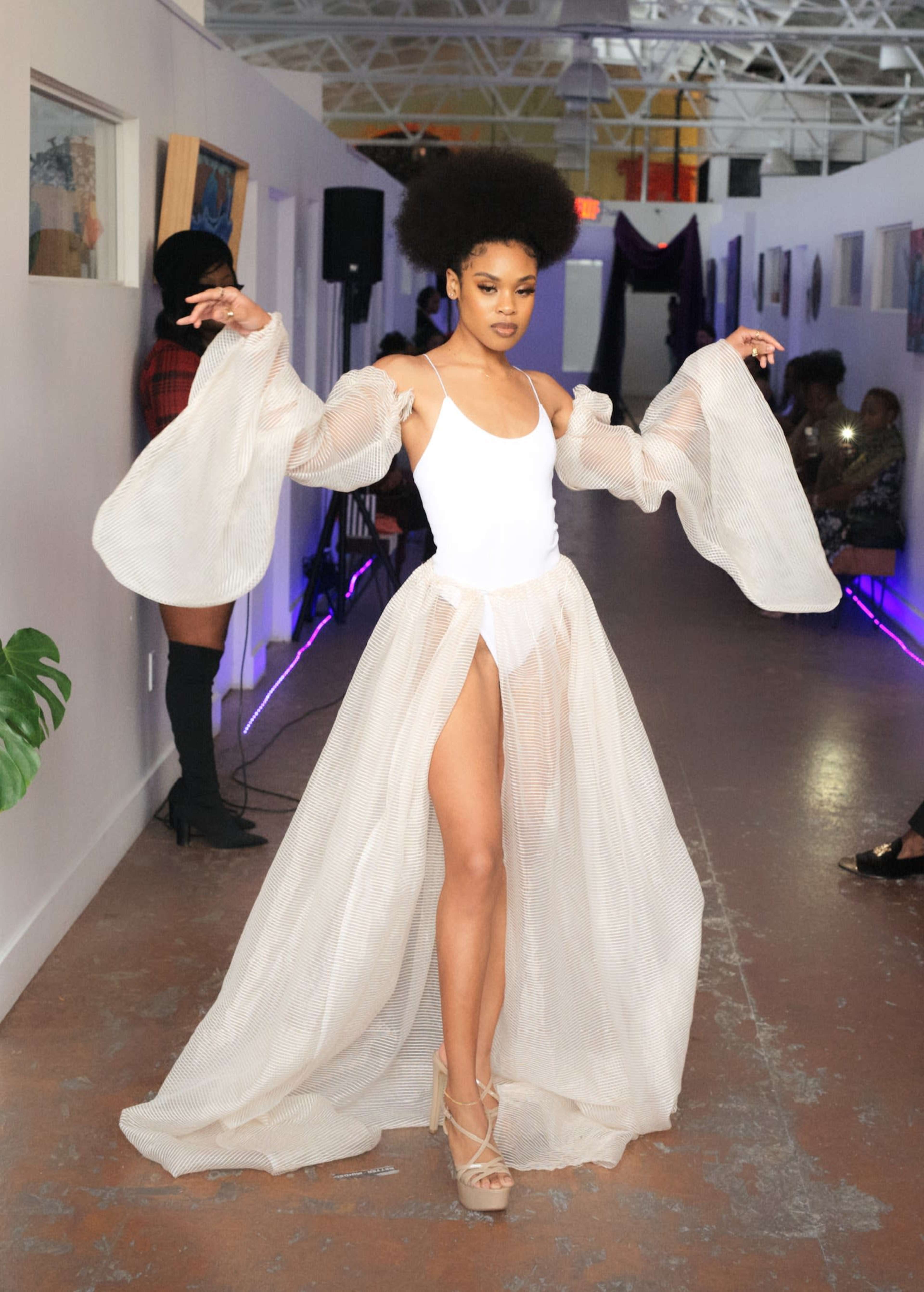A model walks down a narrow hallway wearing a white bodysuit and a flowing, sheer skirt.