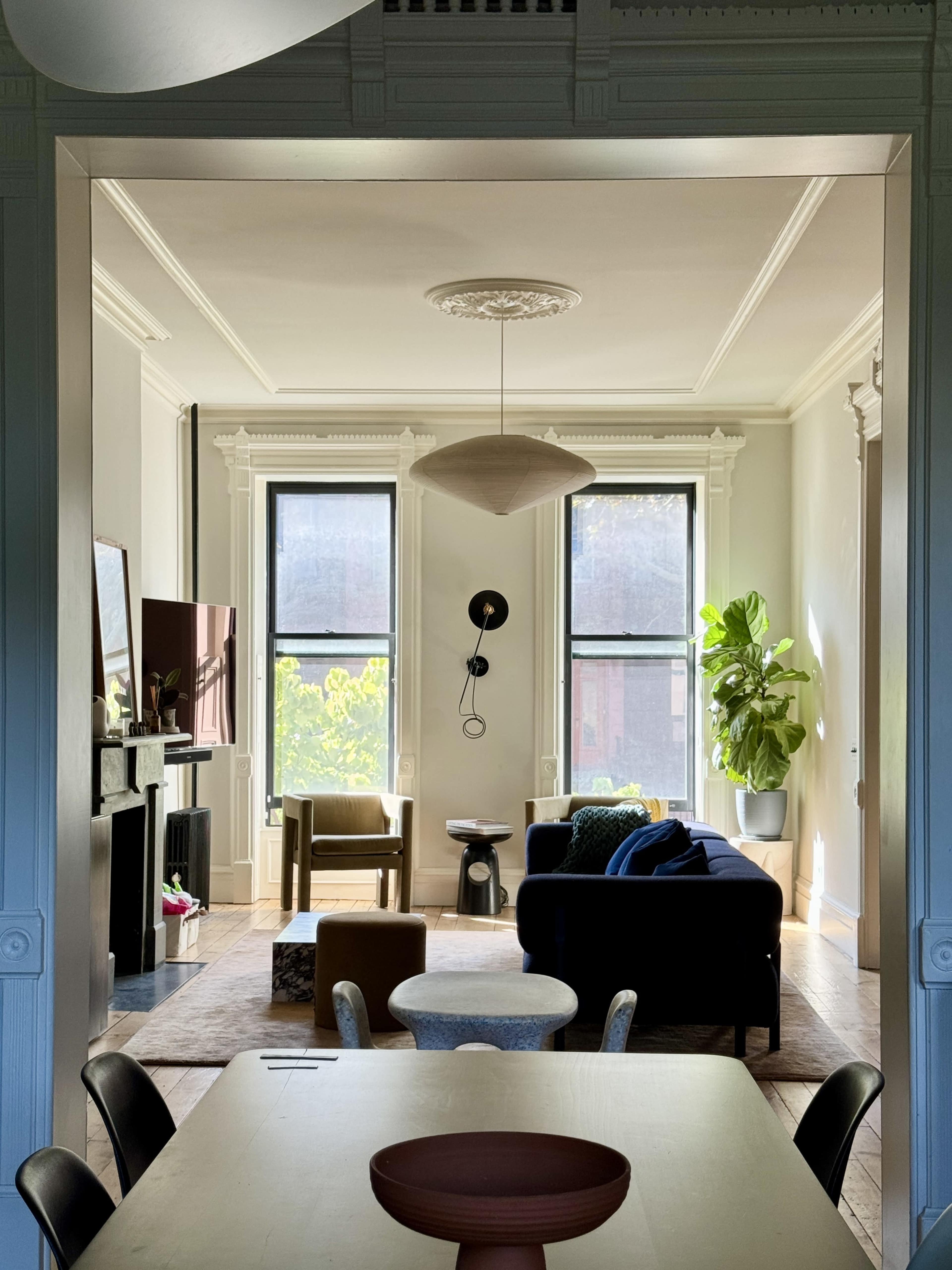 A cozy living room with large windows, a sofa, chairs, and a potted plant, viewed from a dining area.