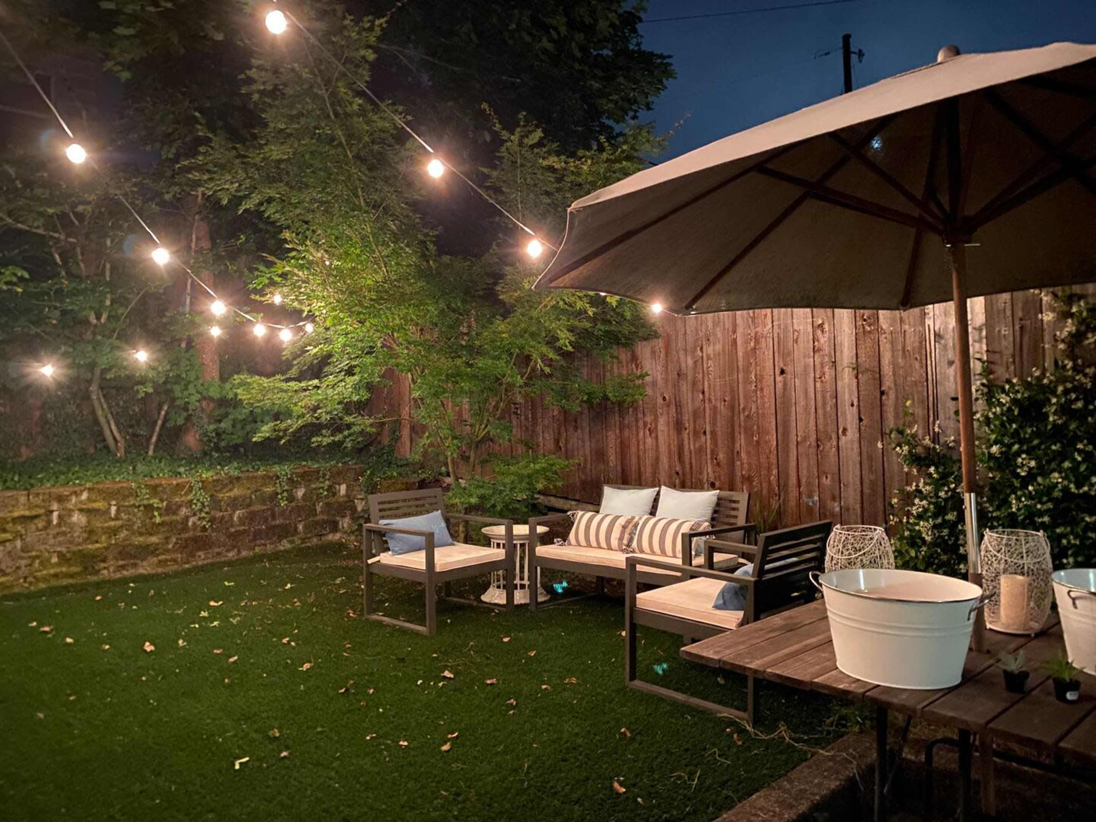 A cozy backyard scene features a seating area with two chairs and a table, surrounded by string lights and greenery.