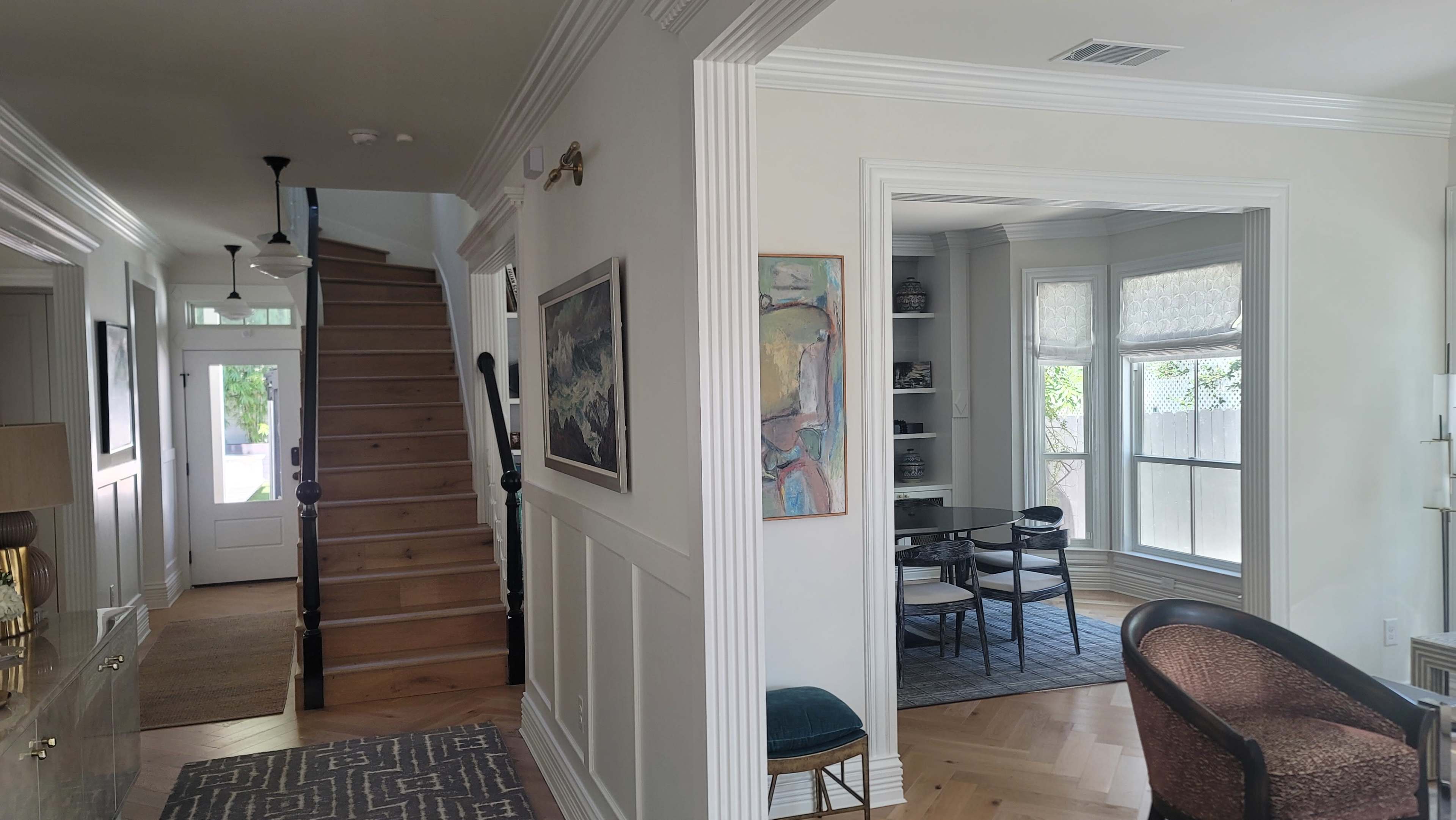 The image shows a bright hallway with a staircase leading up, an adjoining room with large windows, and a small seating area.