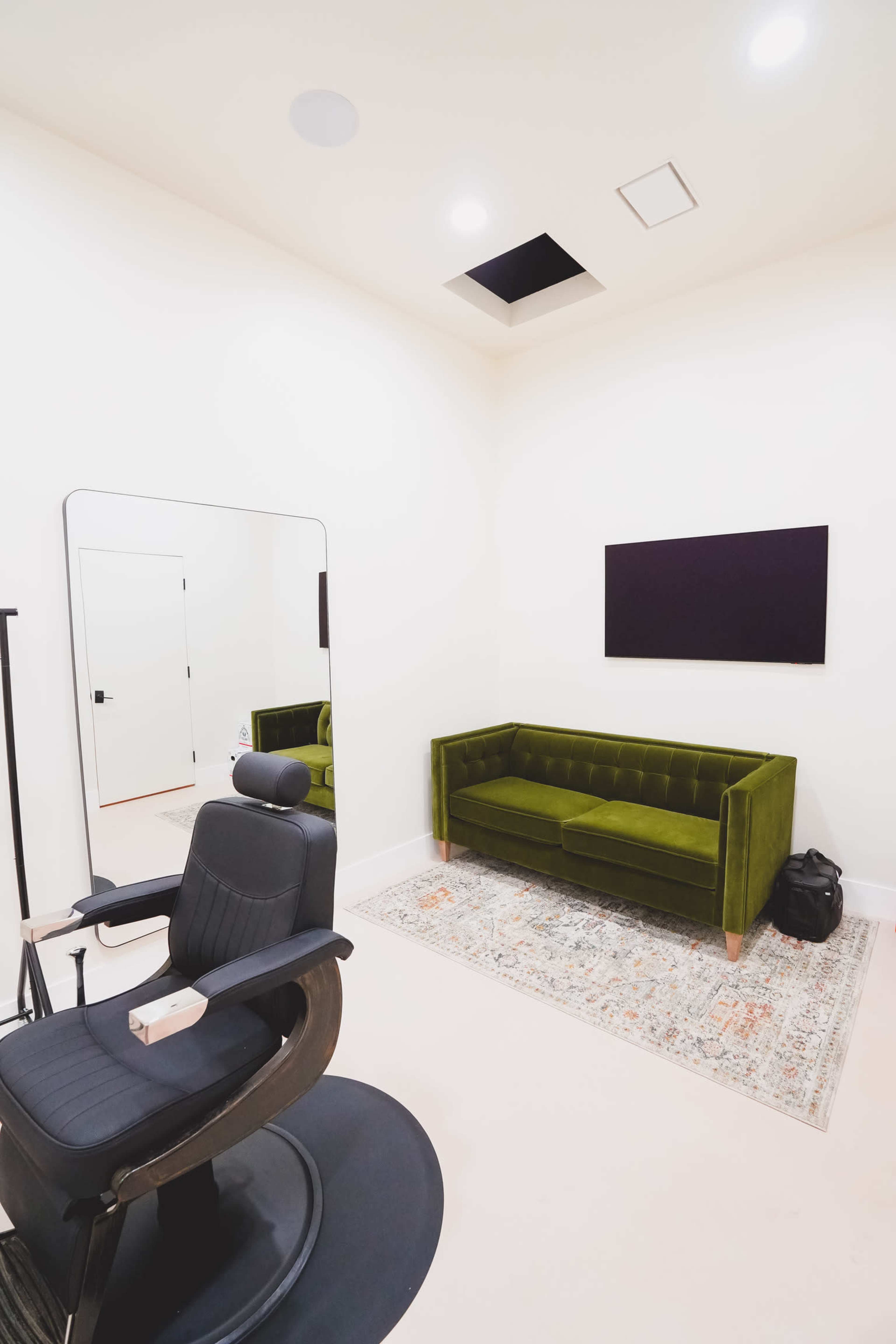 The image shows a modern room featuring a green velvet sofa, a large mirror, a wall-mounted TV, and a black salon chair on a light-colored floor.