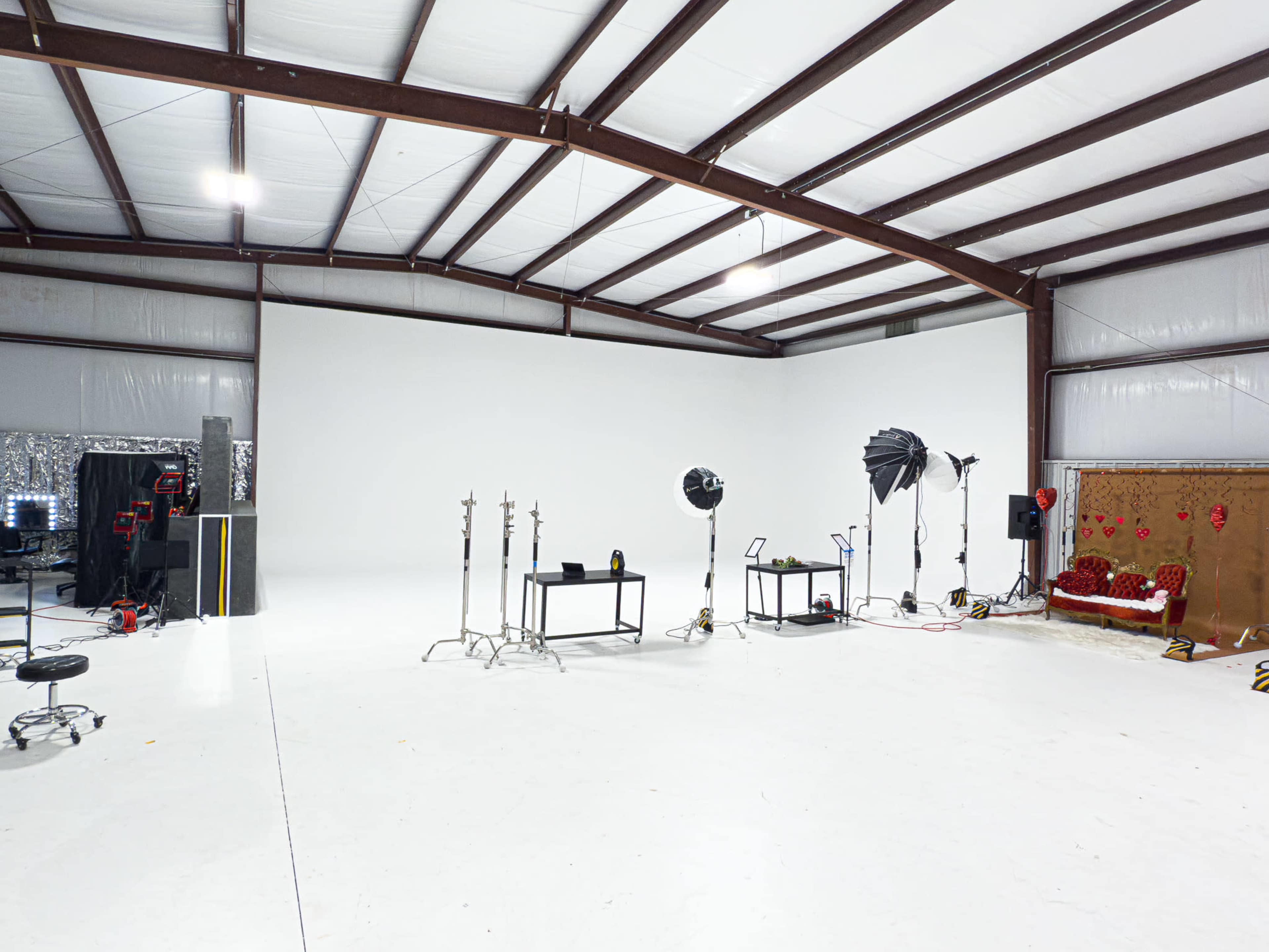 The image shows a spacious, well-lit photography studio featuring a white backdrop, lighting equipment, and various props arranged throughout the area.