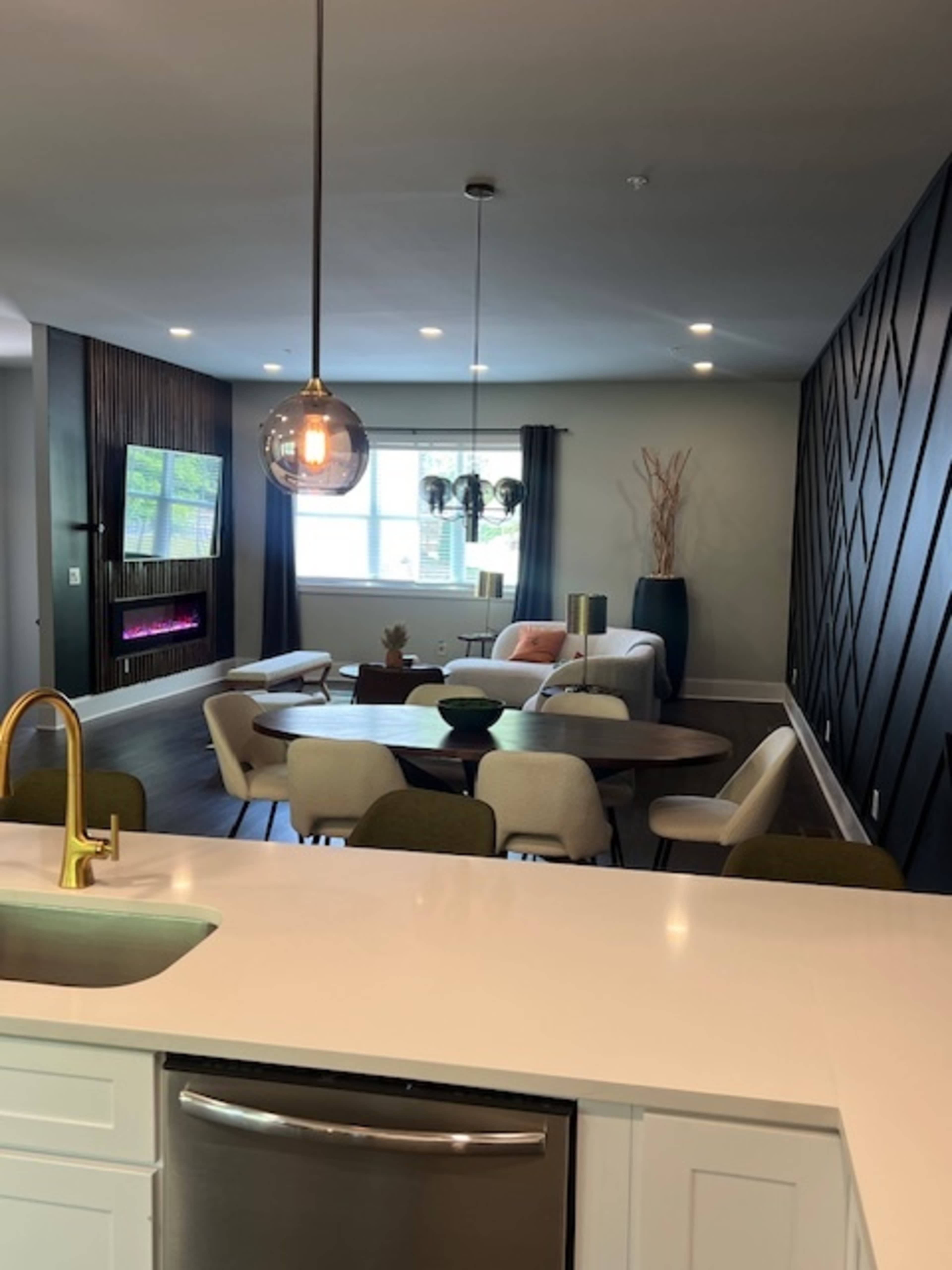 The image shows a modern living space featuring a seating area with a television and a dining table, illuminated by pendant lighting and accented with decorative wall elements.