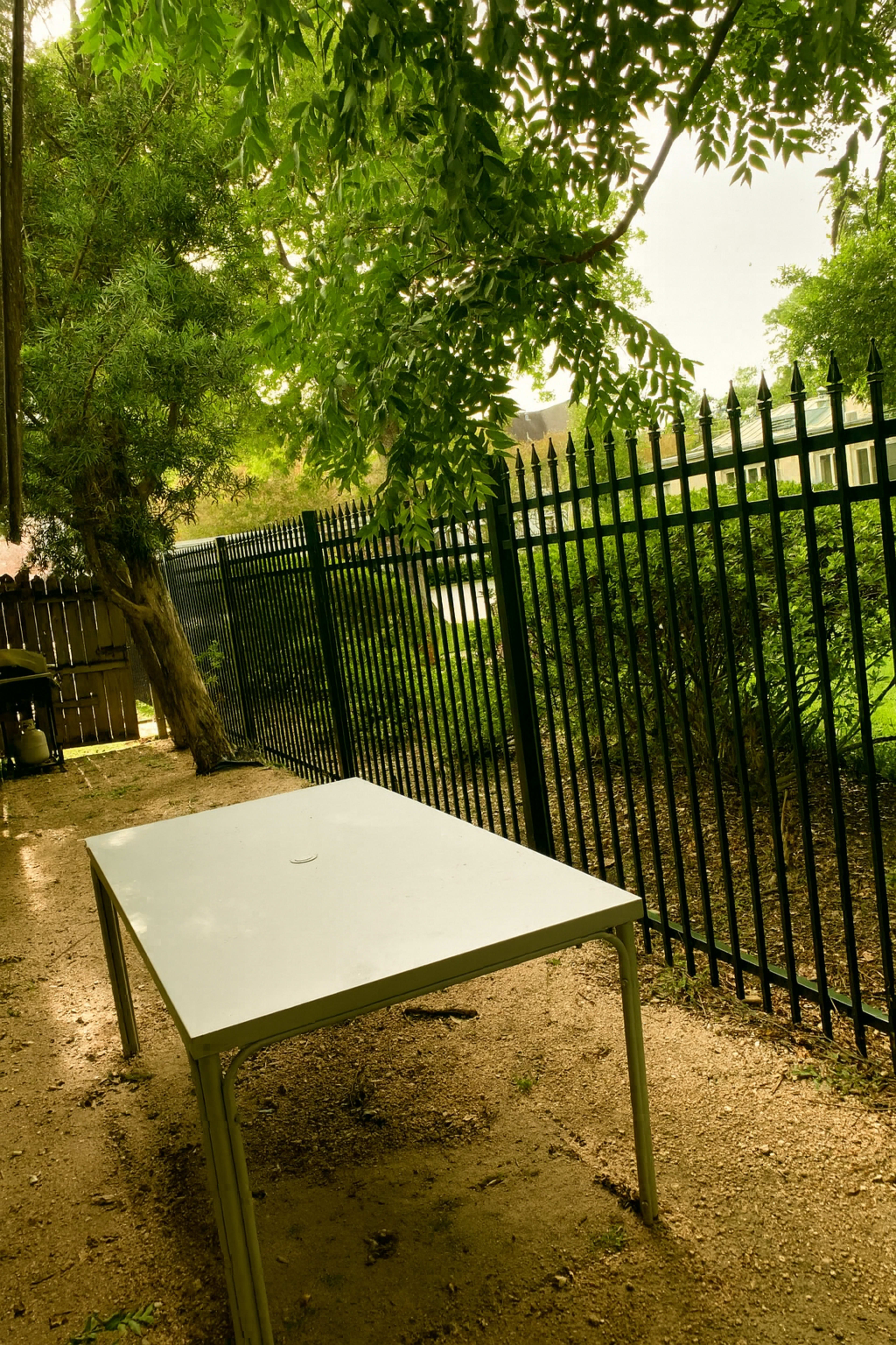 A white table is positioned on a gravel surface in a shaded outdoor area near a black metal fence and surrounded by greenery.