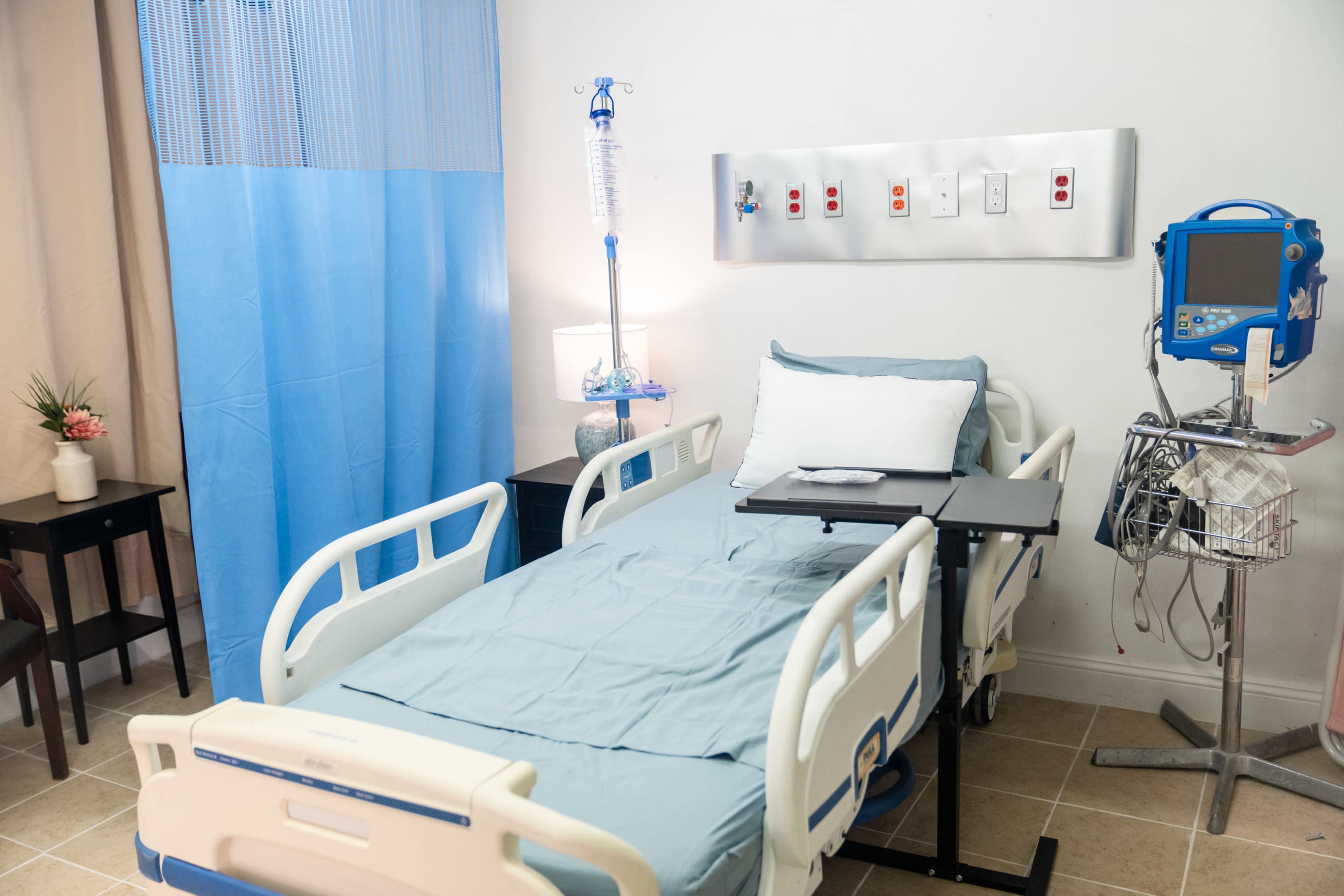 The image shows a hospital room featuring a bed with light blue linens, a bedside table, and various medical equipment along the wall.