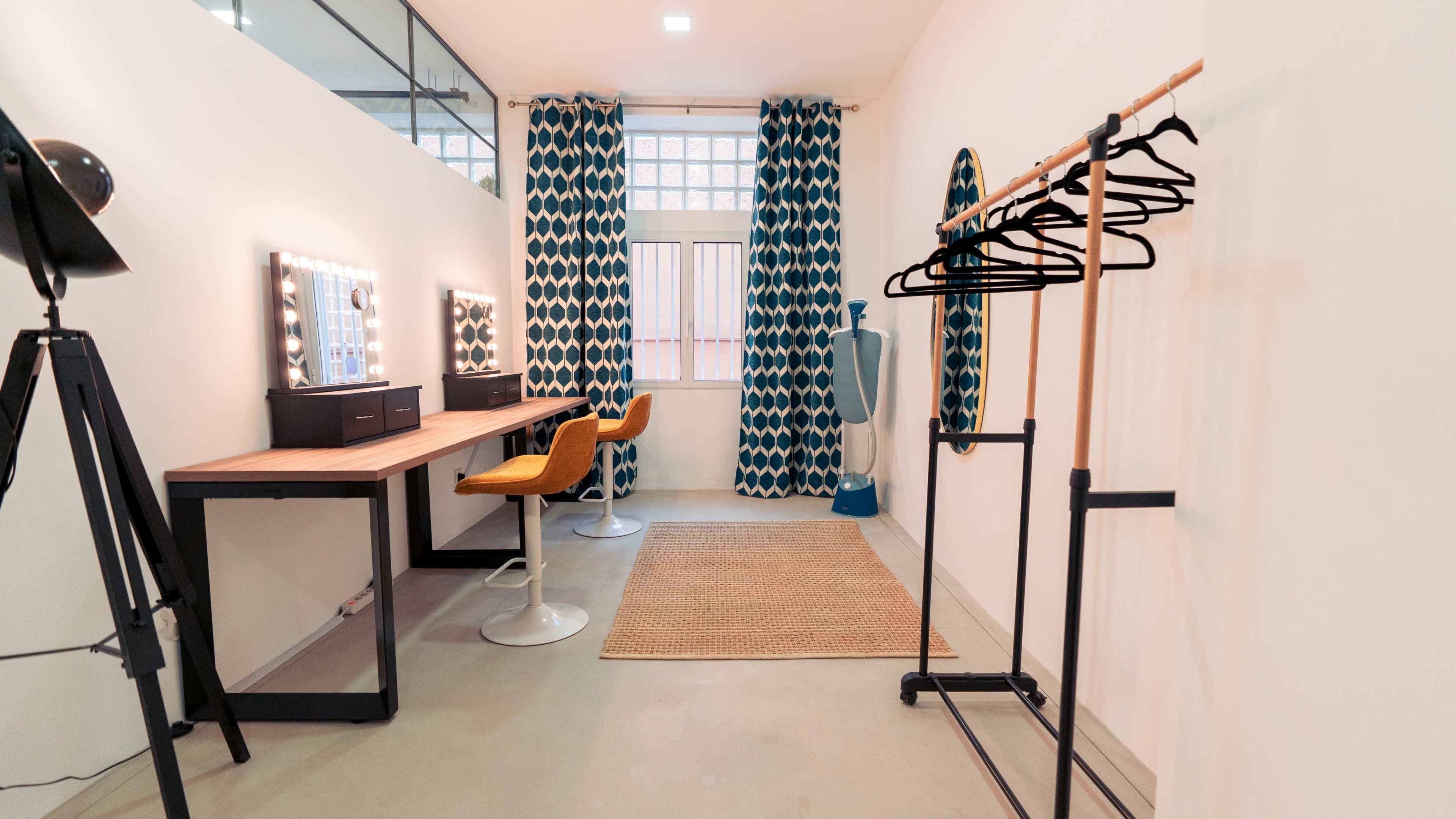 The image shows a bright room with a dressing area featuring a mirror with lights, a desk, an orange chair, a clothing rack, and patterned curtains.