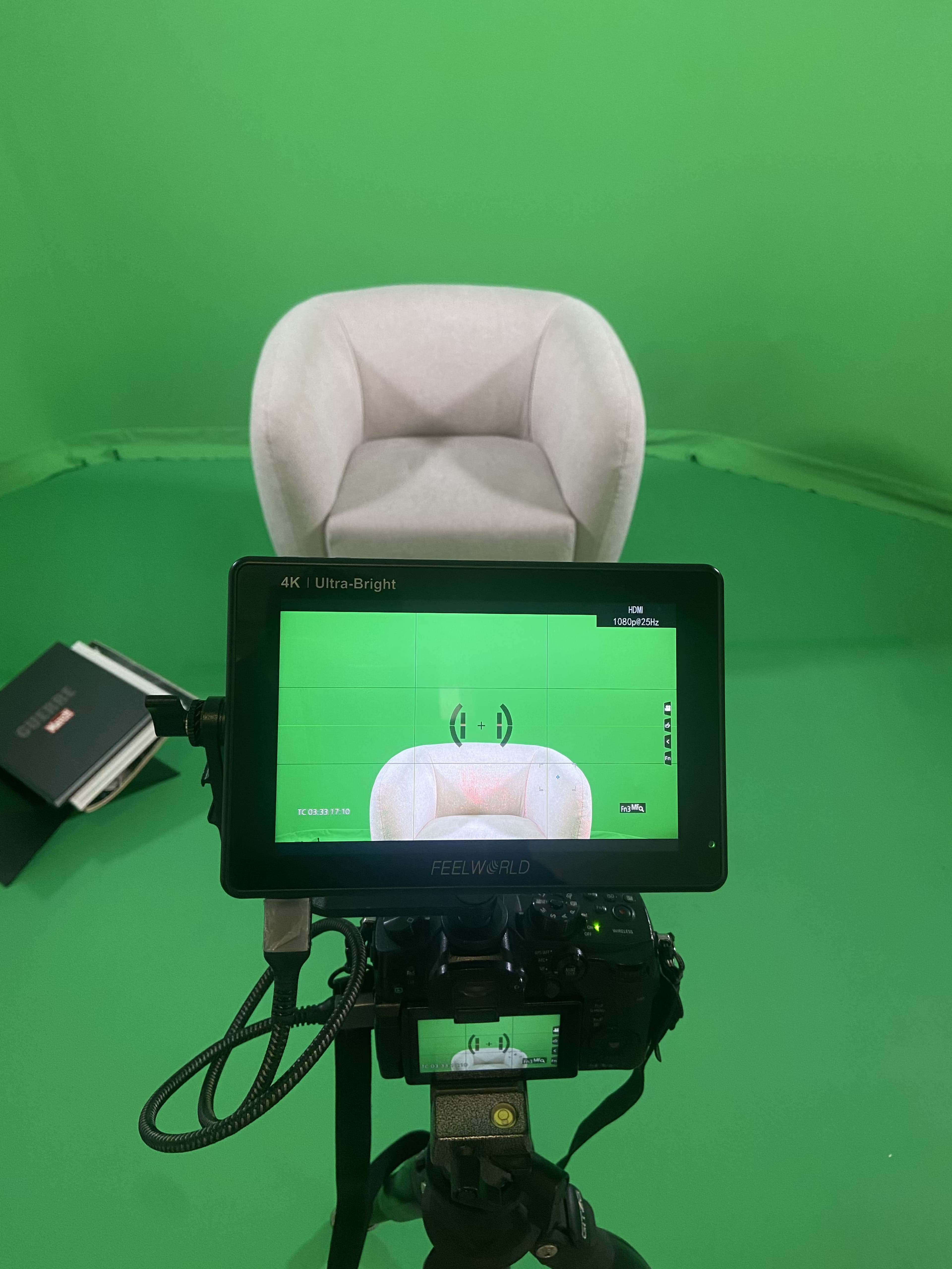 A camera and monitor are positioned in front of a white chair against a green screen backdrop.