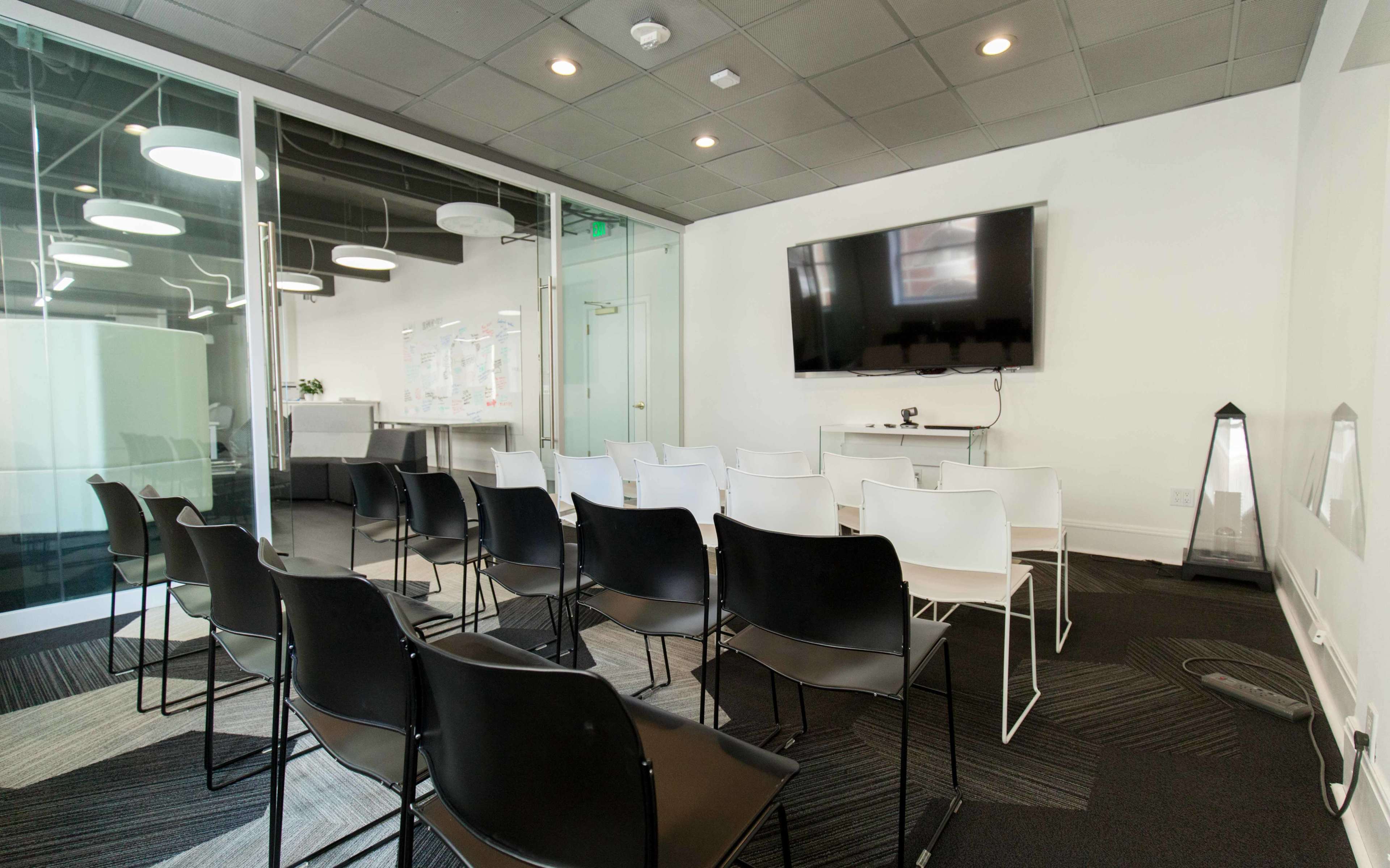 A modern meeting room with rows of black and white chairs facing a large mounted television screen.