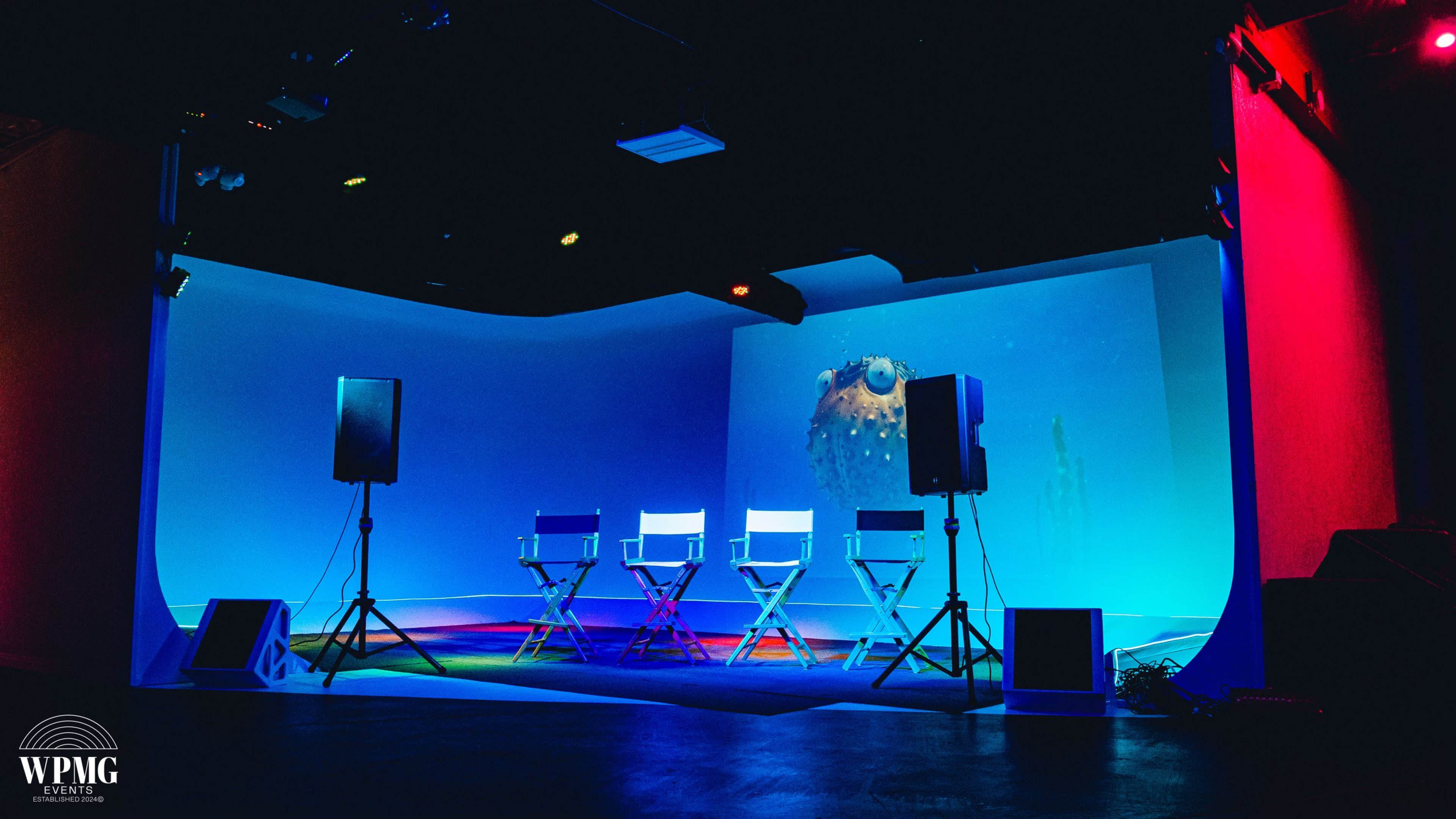 A minimalist stage setup features four empty director's chairs in front of a large screen, illuminated by colorful lights.