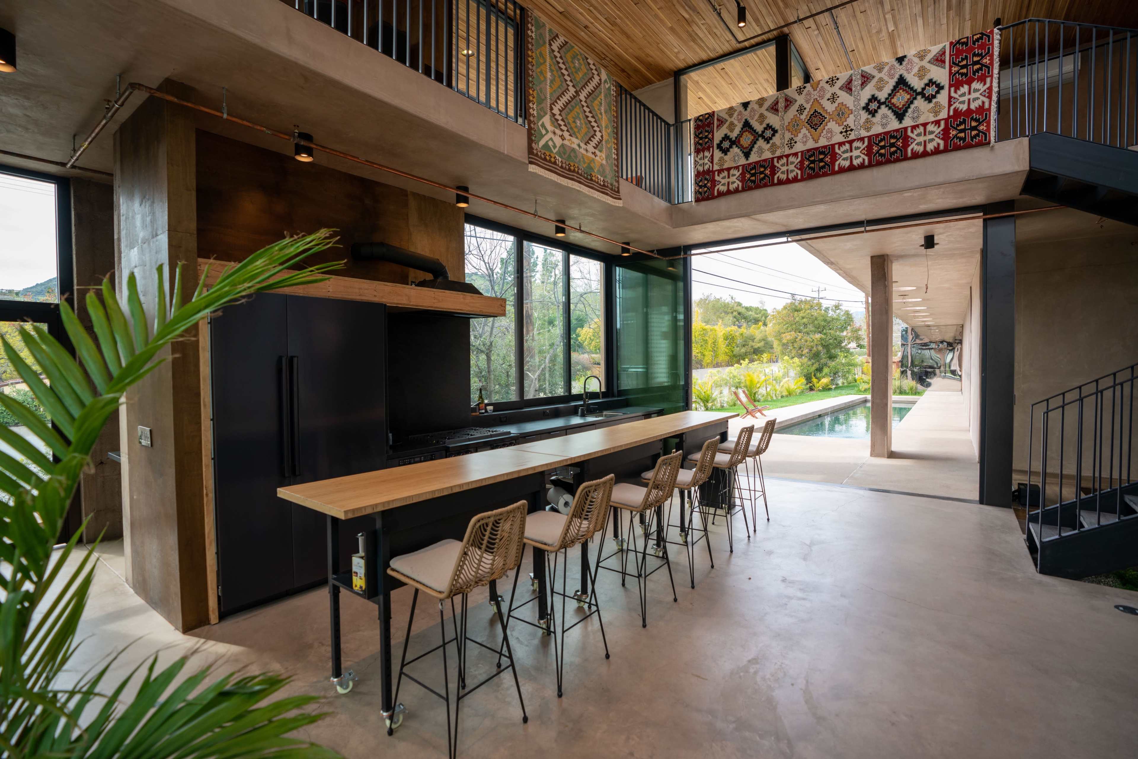 The image shows a modern kitchen with a long wooden bar counter and stools, large windows overlooking a garden, and a staircase leading to another level.