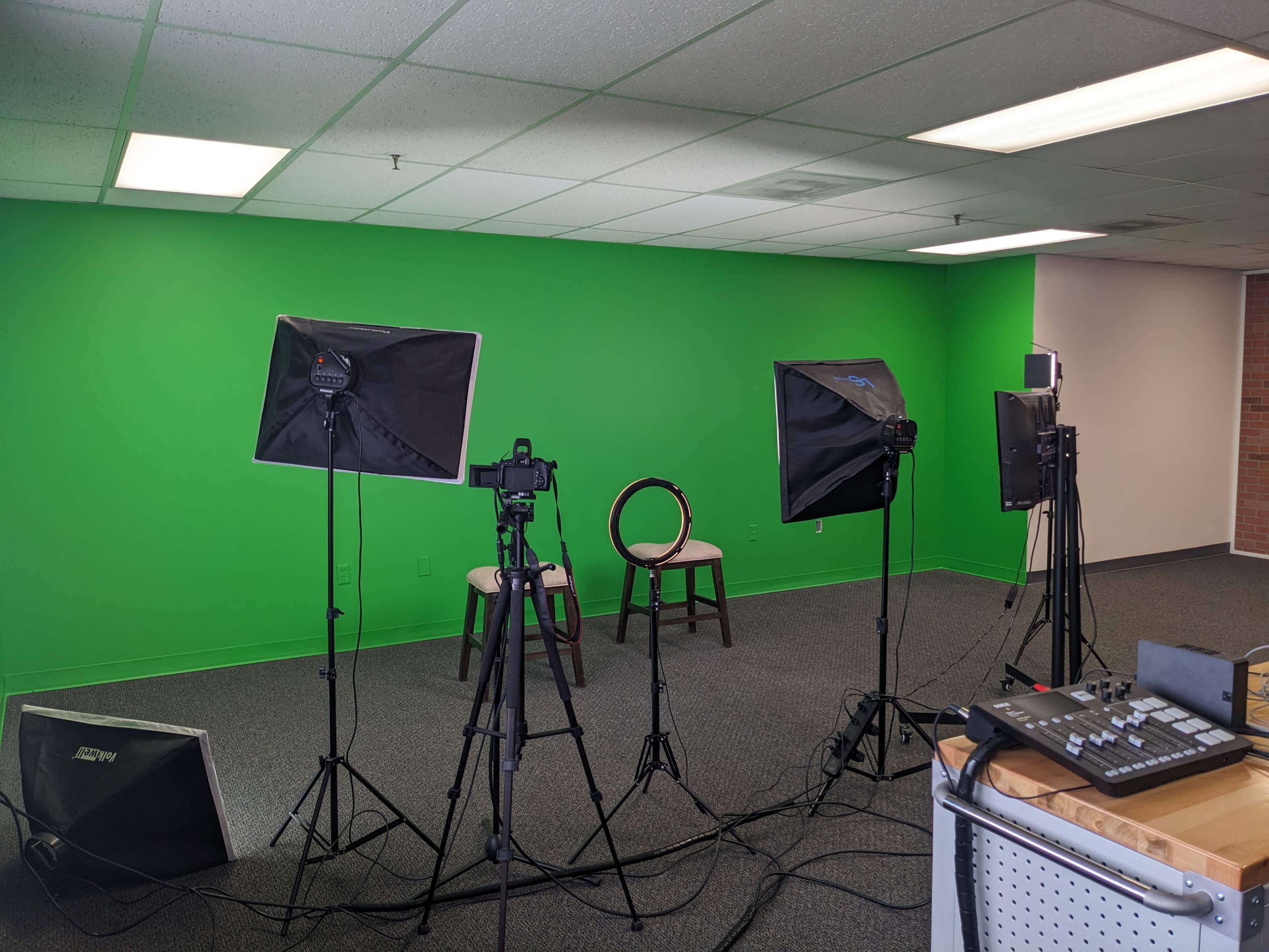 The image shows a studio setup with three tripods holding lights and cameras, directed towards a pair of chairs in front of a bright green wall.