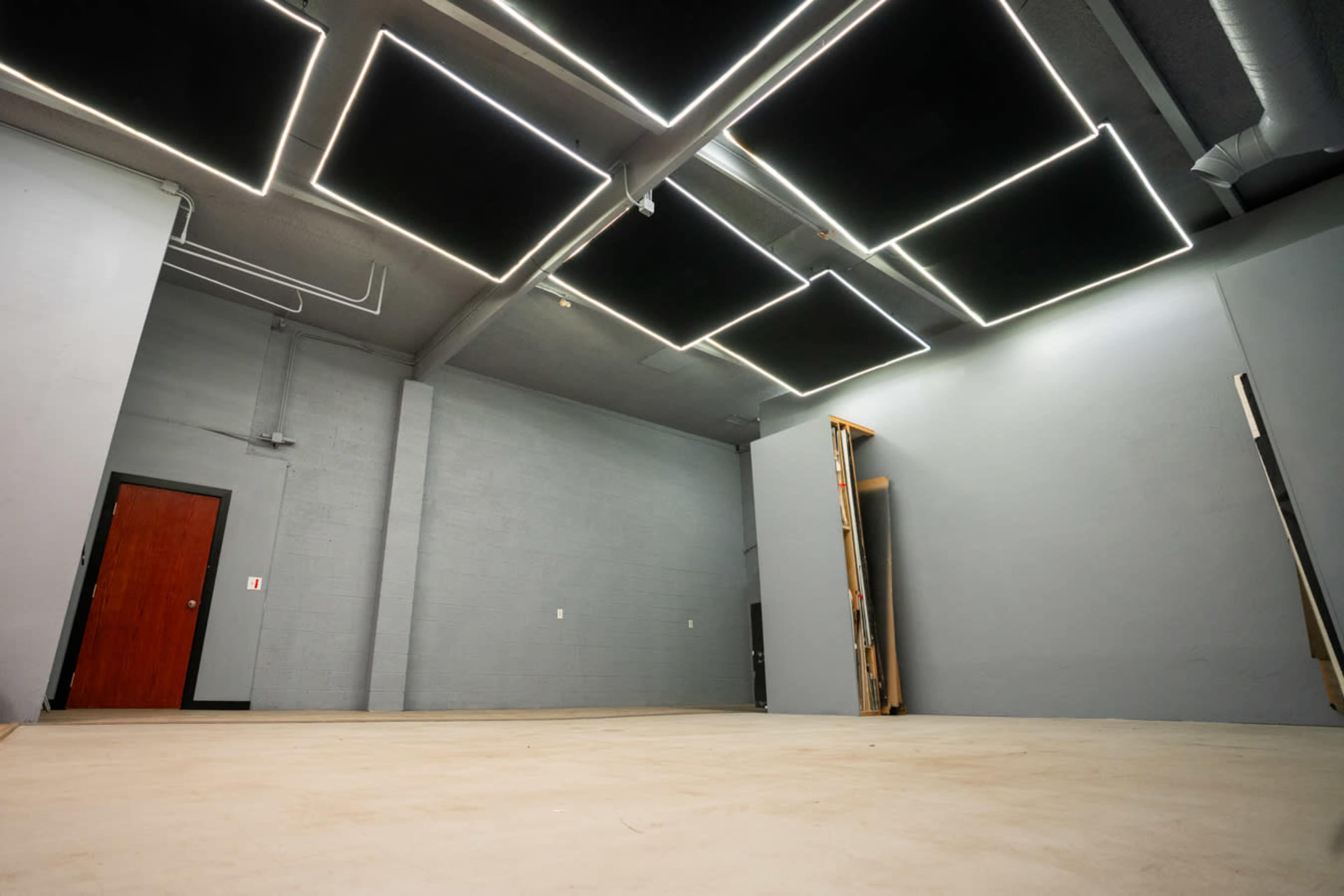 The image shows an empty industrial-style room with gray walls, a concrete floor, and a ceiling featuring rectangular light panels.