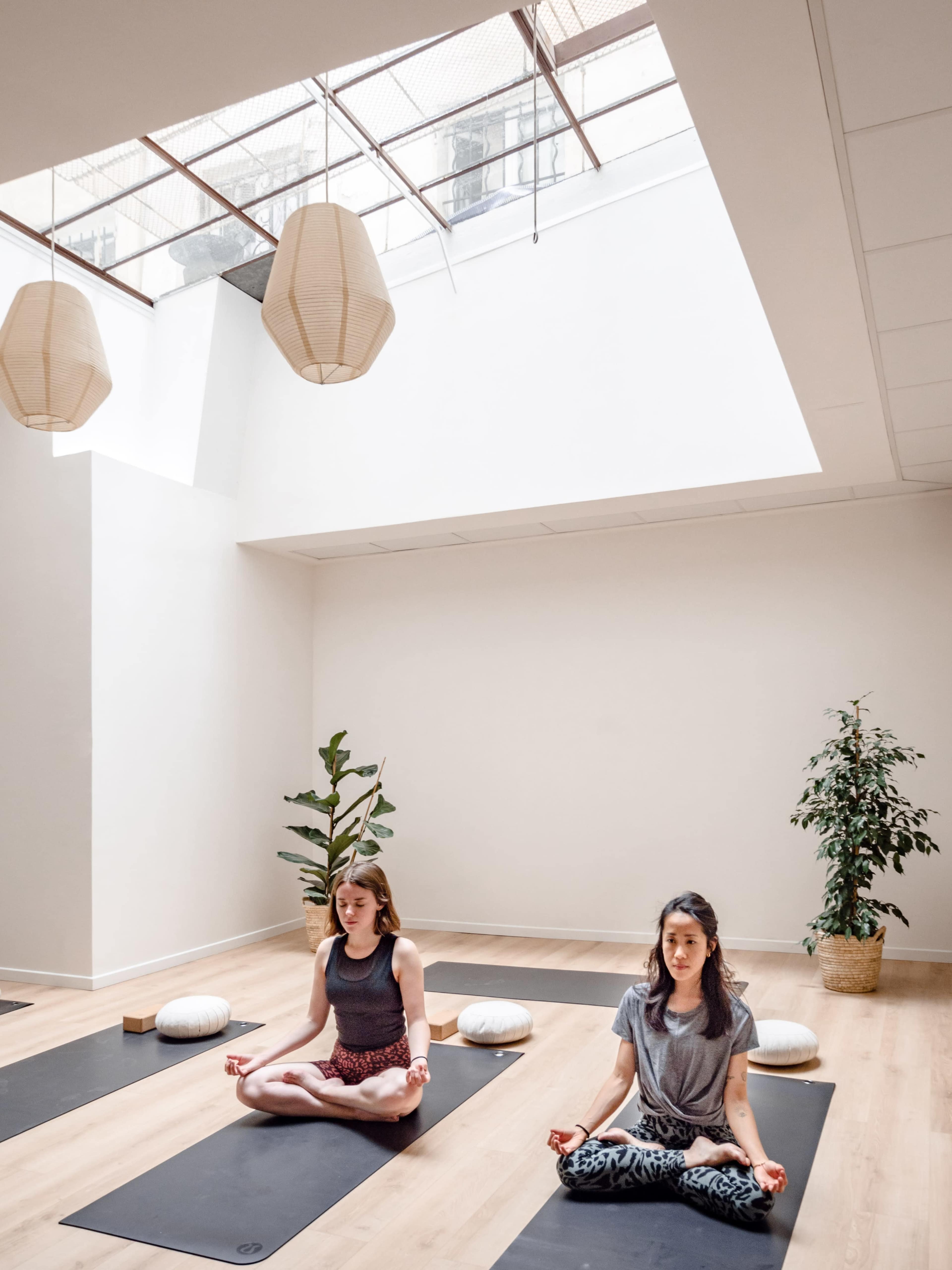 Two women practice yoga in a spacious studio with natural light, sitting on black mats surrounded by plants and meditation cushions.
