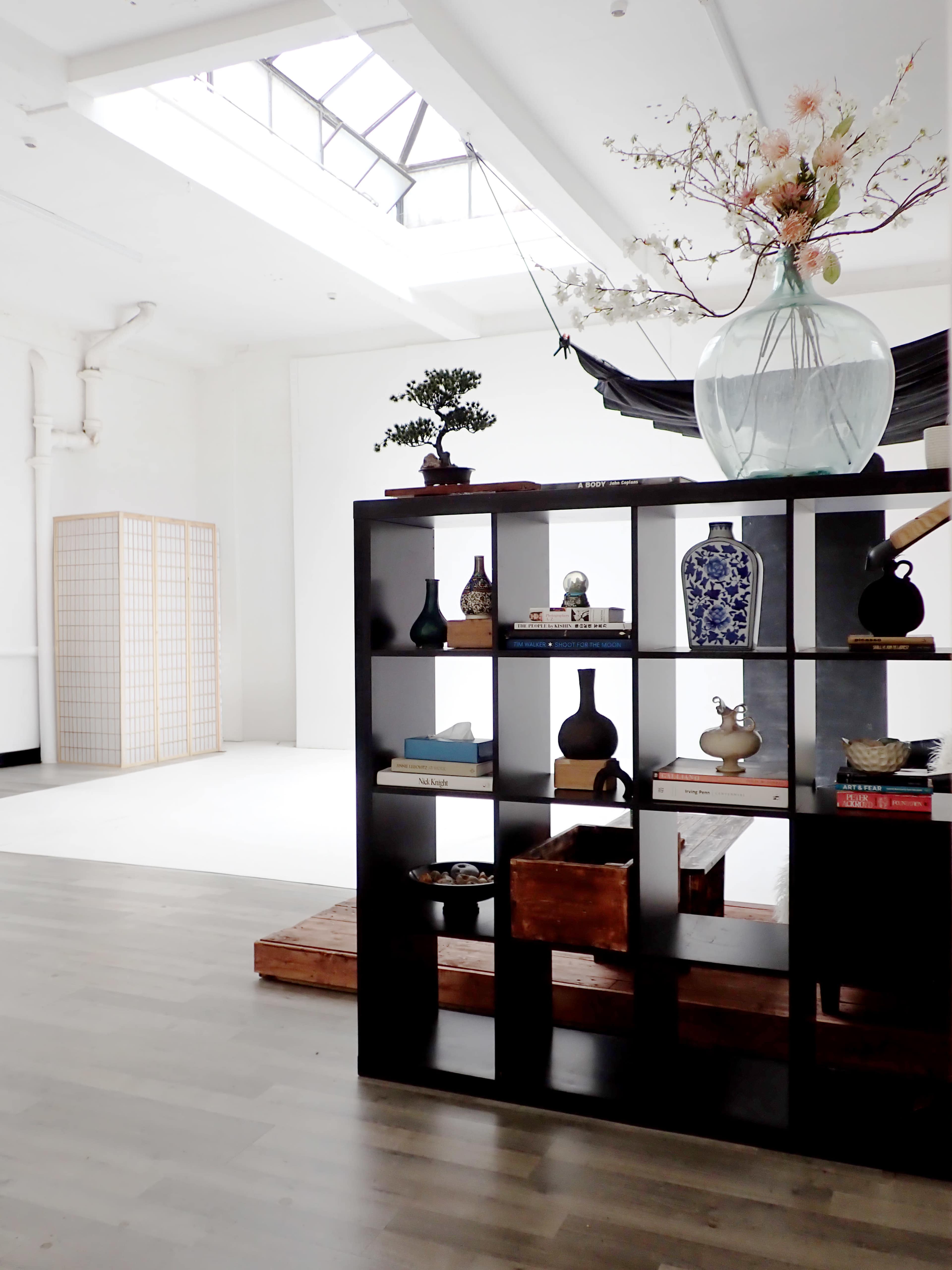 The image shows a spacious, minimalist interior with a black bookshelf containing decorative items and a large vase with flowers, while natural light enters through a skylight.