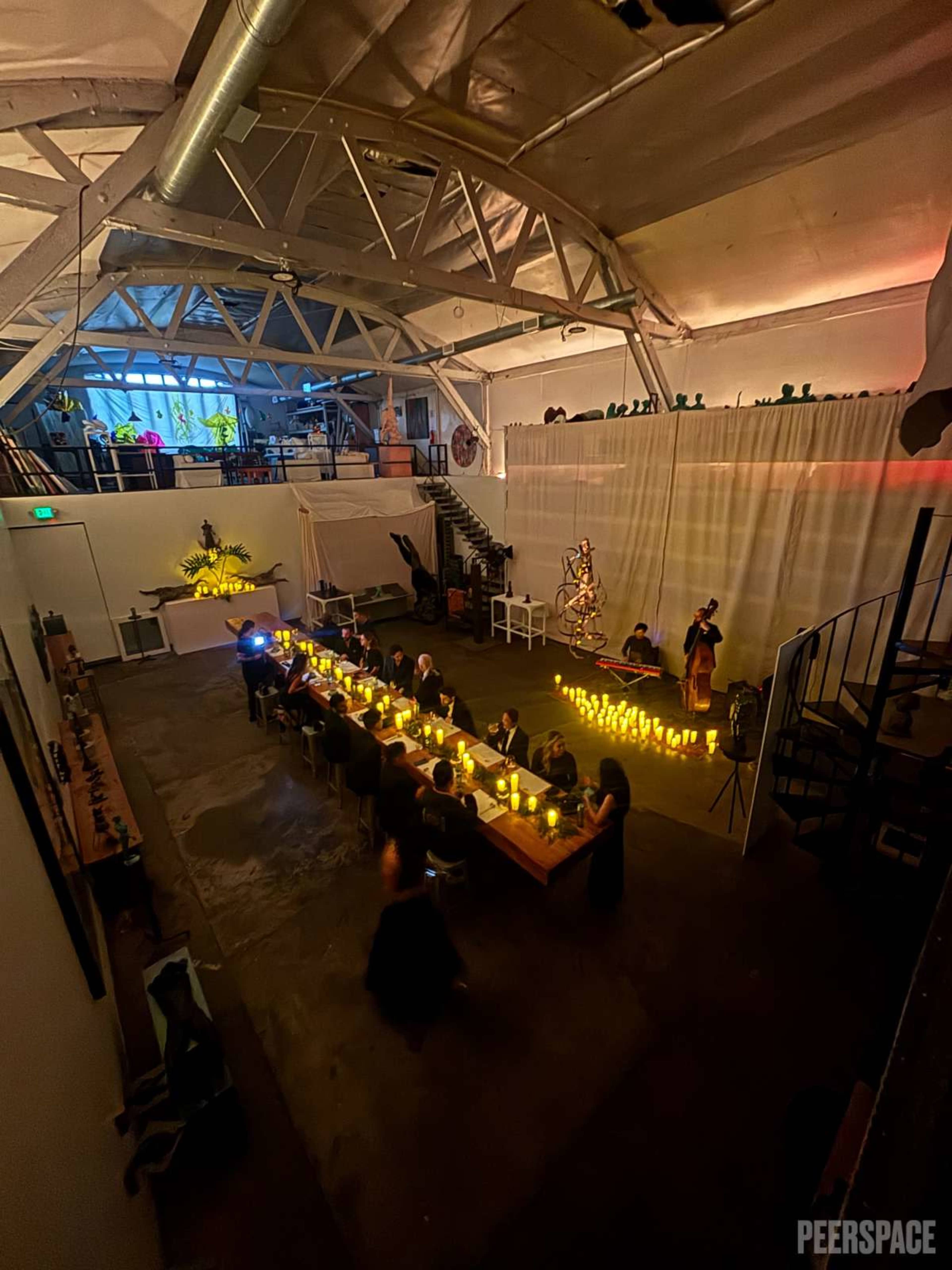 A long table set with candles occupies a spacious, dimly lit event space featuring a staircase and decorative elements in the background.