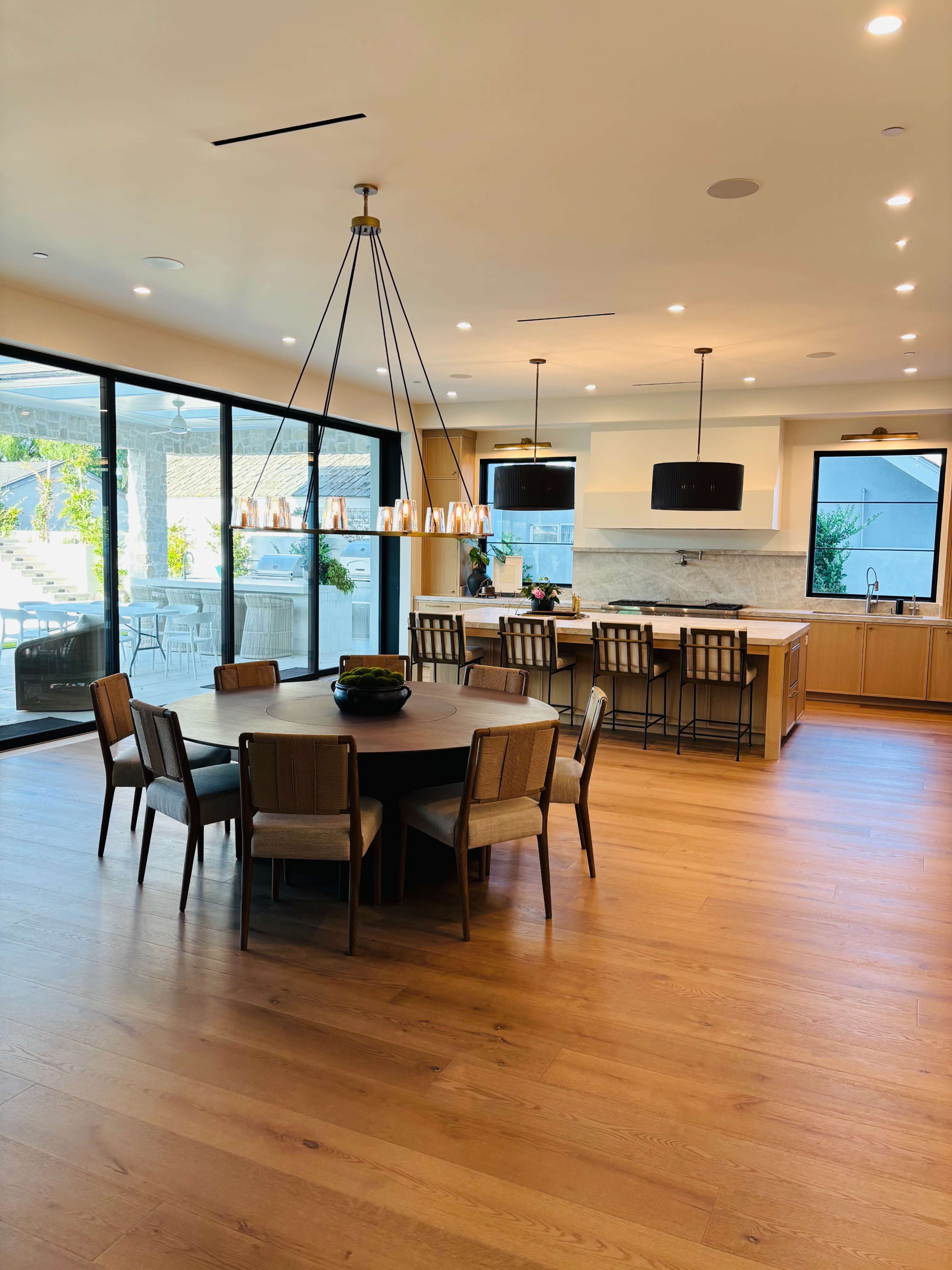 The image shows a modern kitchen and dining area with a round wooden table surrounded by upholstered chairs, large glass doors leading to an outdoor space, and a contemporary light fixture overhead.