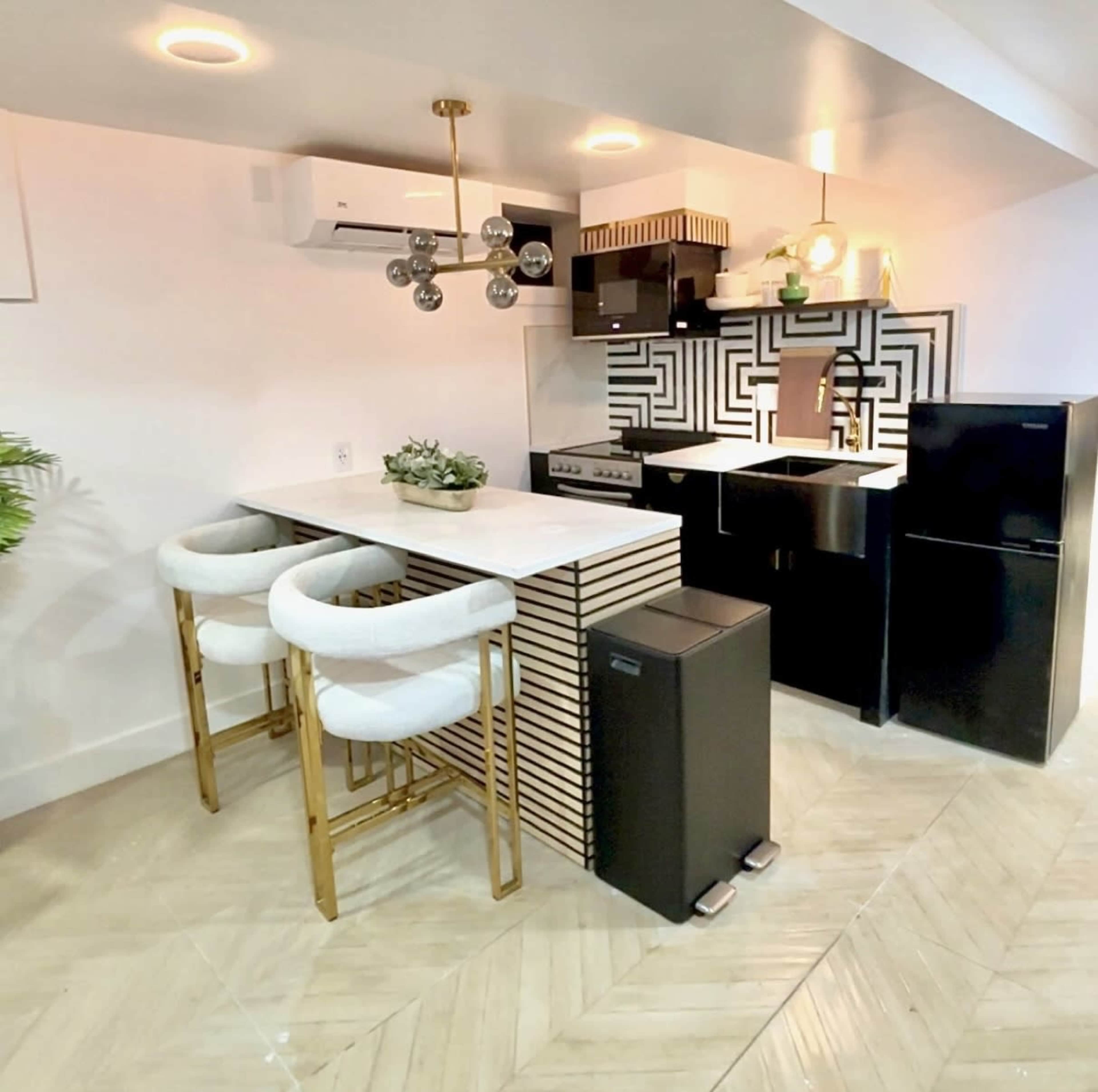 The image shows a modern kitchen with black and white cabinets, a marble countertop, and bar stools with a gold frame.