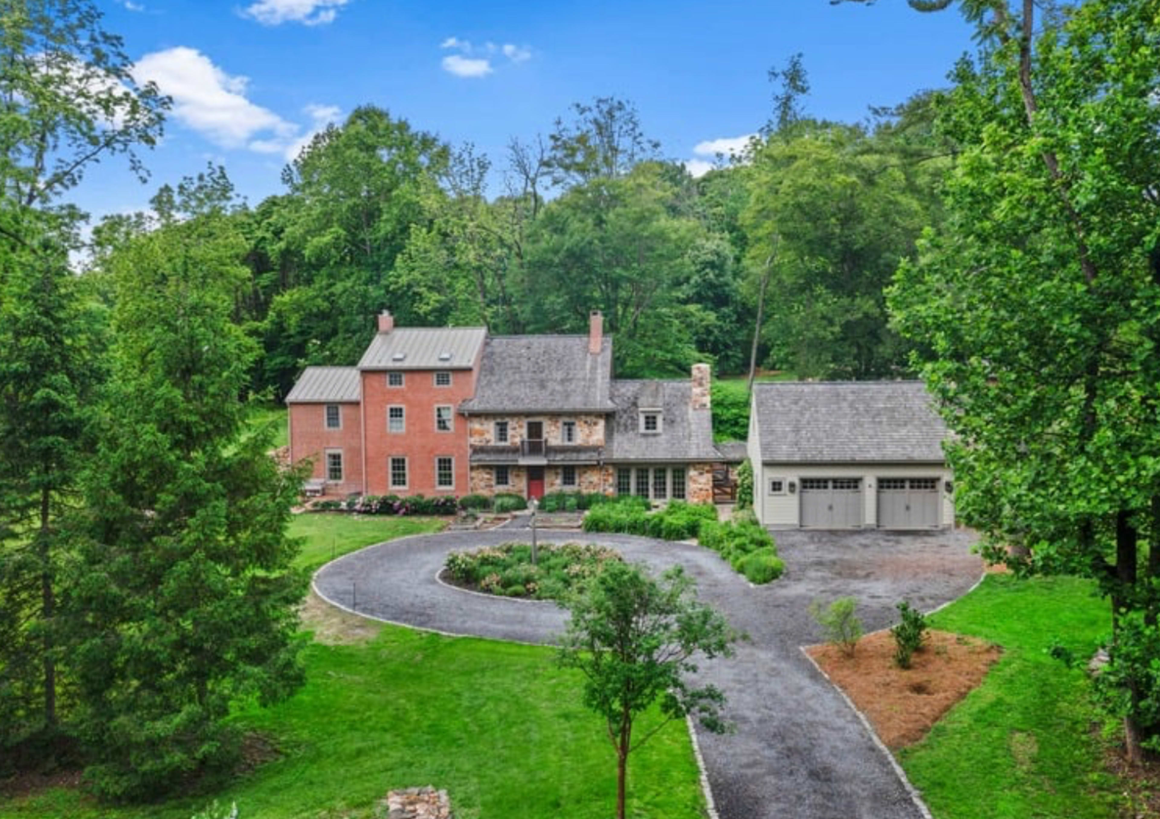 The image shows a large, two-story house with a blend of brick and wood exteriors, situated on a landscaped property surrounded by trees and a gravel circular driveway.