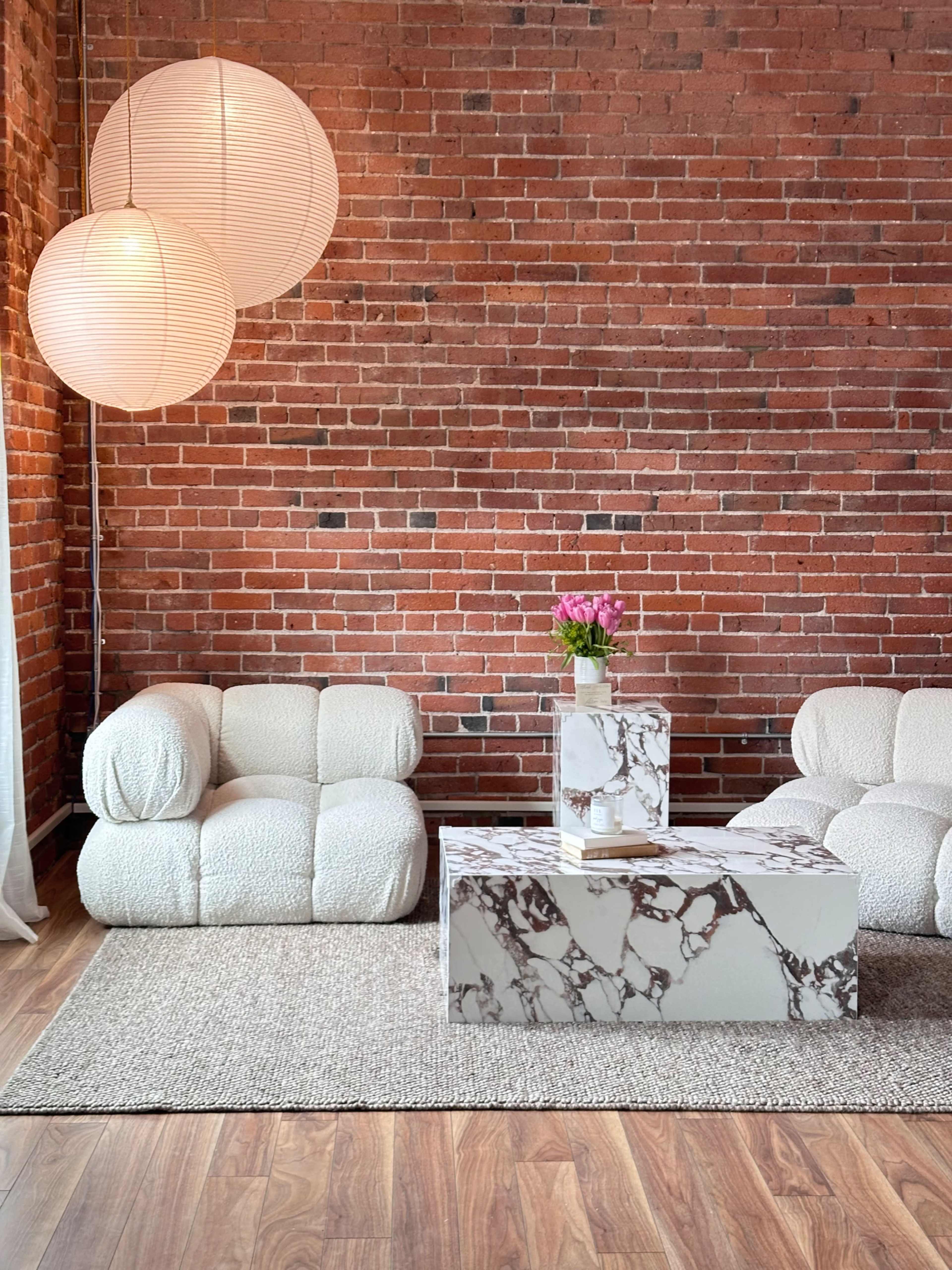 The image features a cozy living room with two soft, textured sofas, a marble coffee table, and two large hanging light fixtures against a backdrop of exposed brick walls.