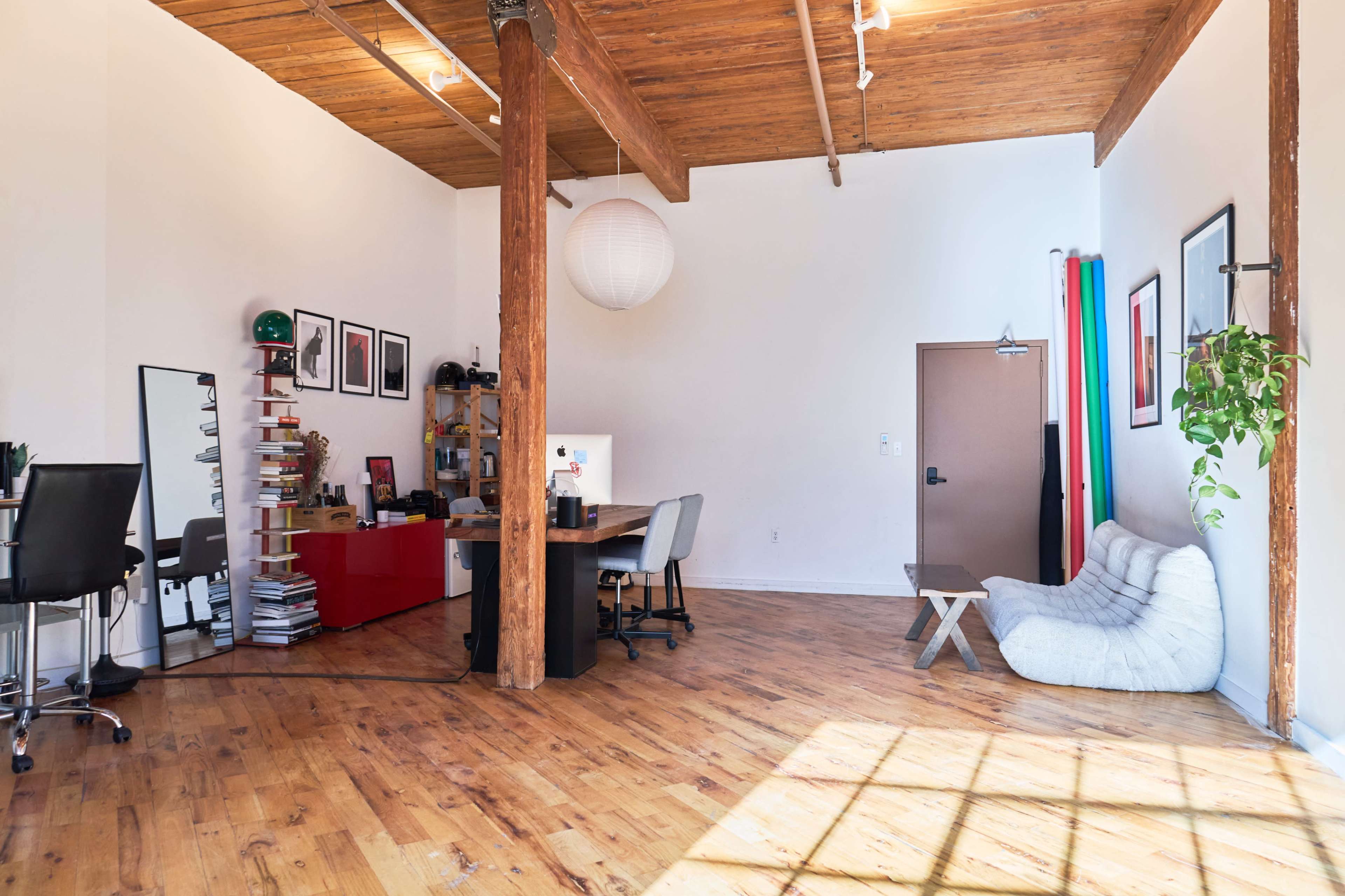 The image shows a spacious, well-lit office featuring wooden floors, exposed beams, a desk with a computer, and a cozy seating area.