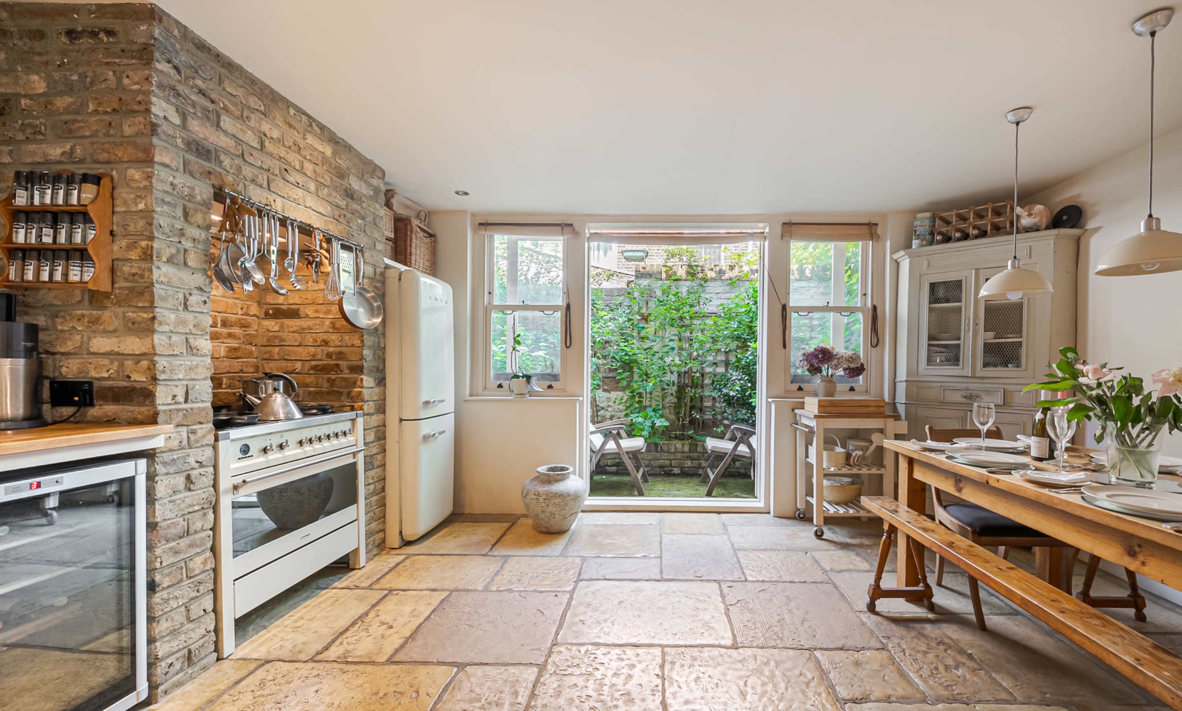 The kitchen features exposed brick walls, a large window overlooking a garden, and a wooden dining table.