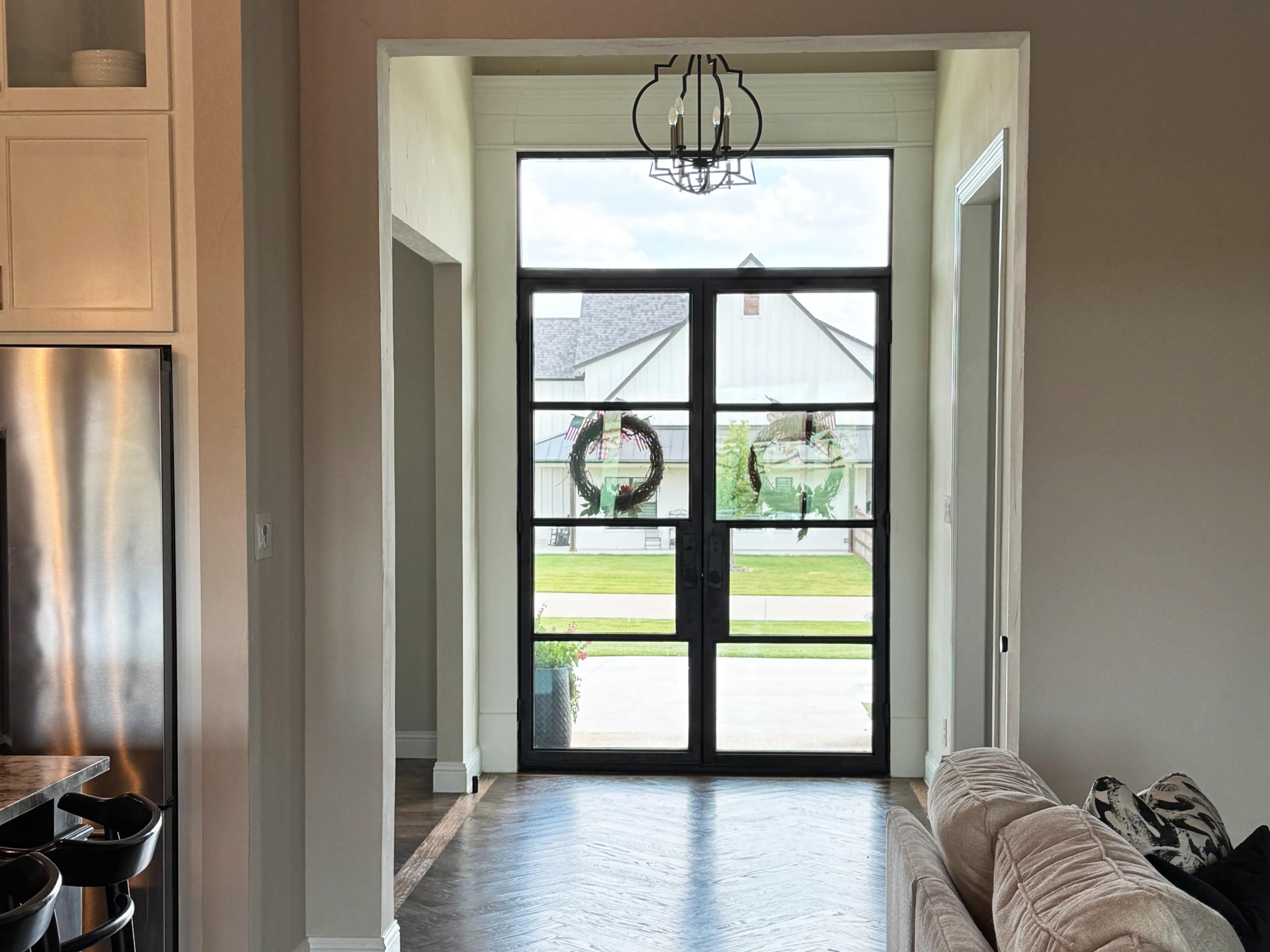 The image shows an entrance area with a large glass door framed by a light-colored wall, leading to an outdoor space with greenery visible through the door.