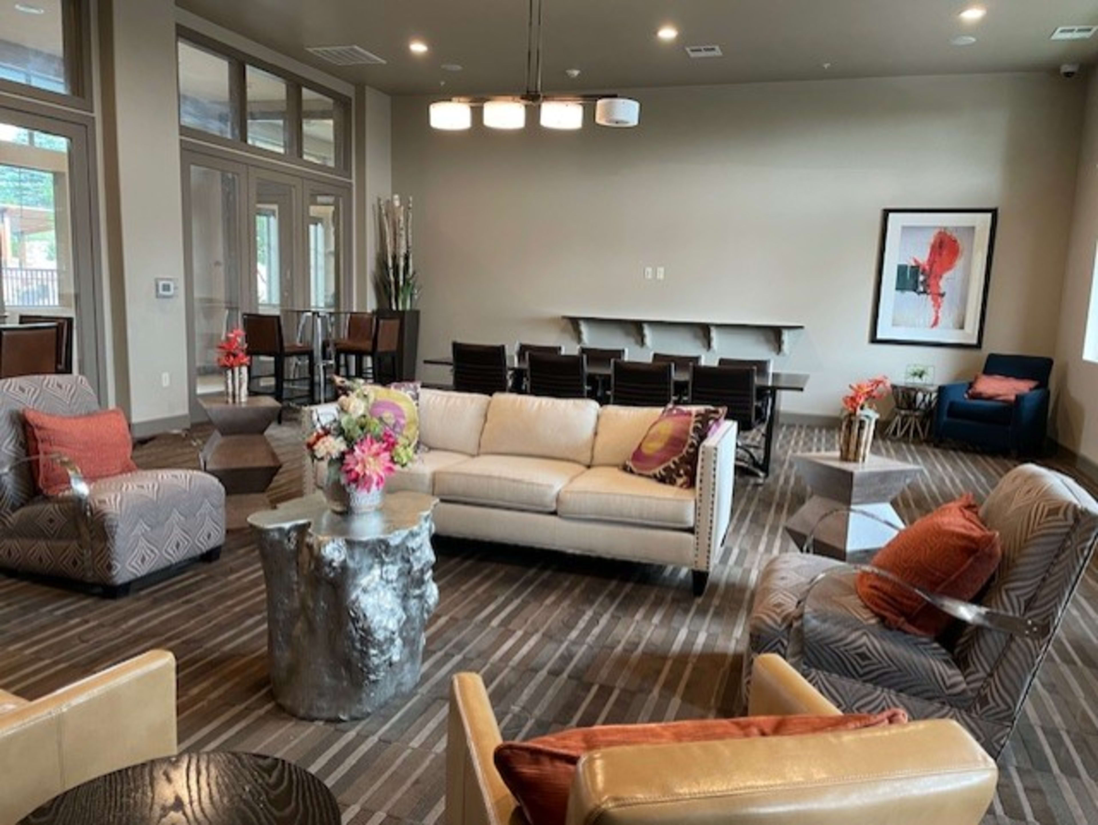 The image shows a modern lounge area featuring a beige sofa, several patterned chairs, a large floral centerpiece on a side table, and a dining space in the background.