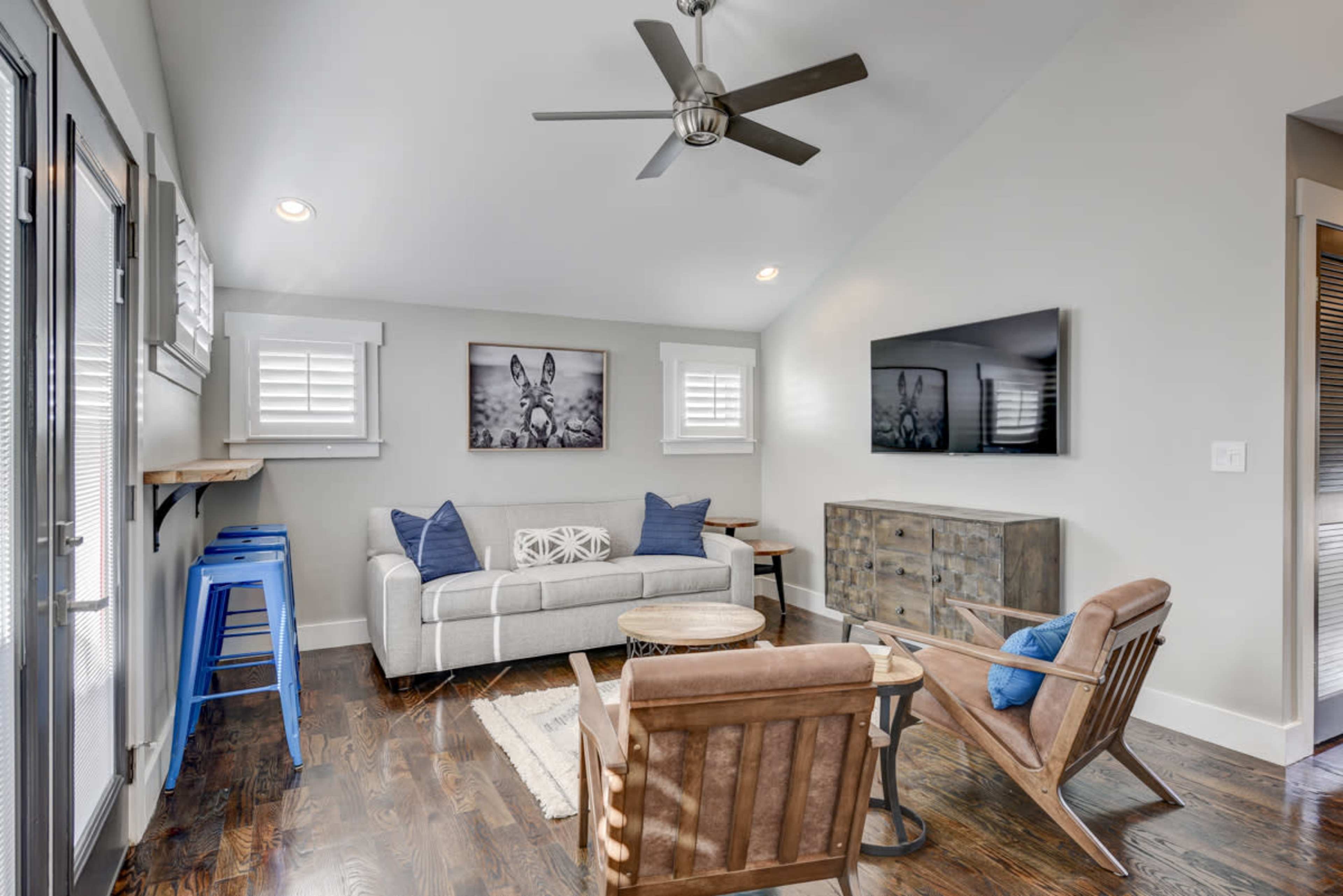 A modern living room featuring a light gray sofa, two wooden armchairs, a round coffee table, and a mounted television on the wall, with a ceiling fan overhead and decorative artwork displayed.
