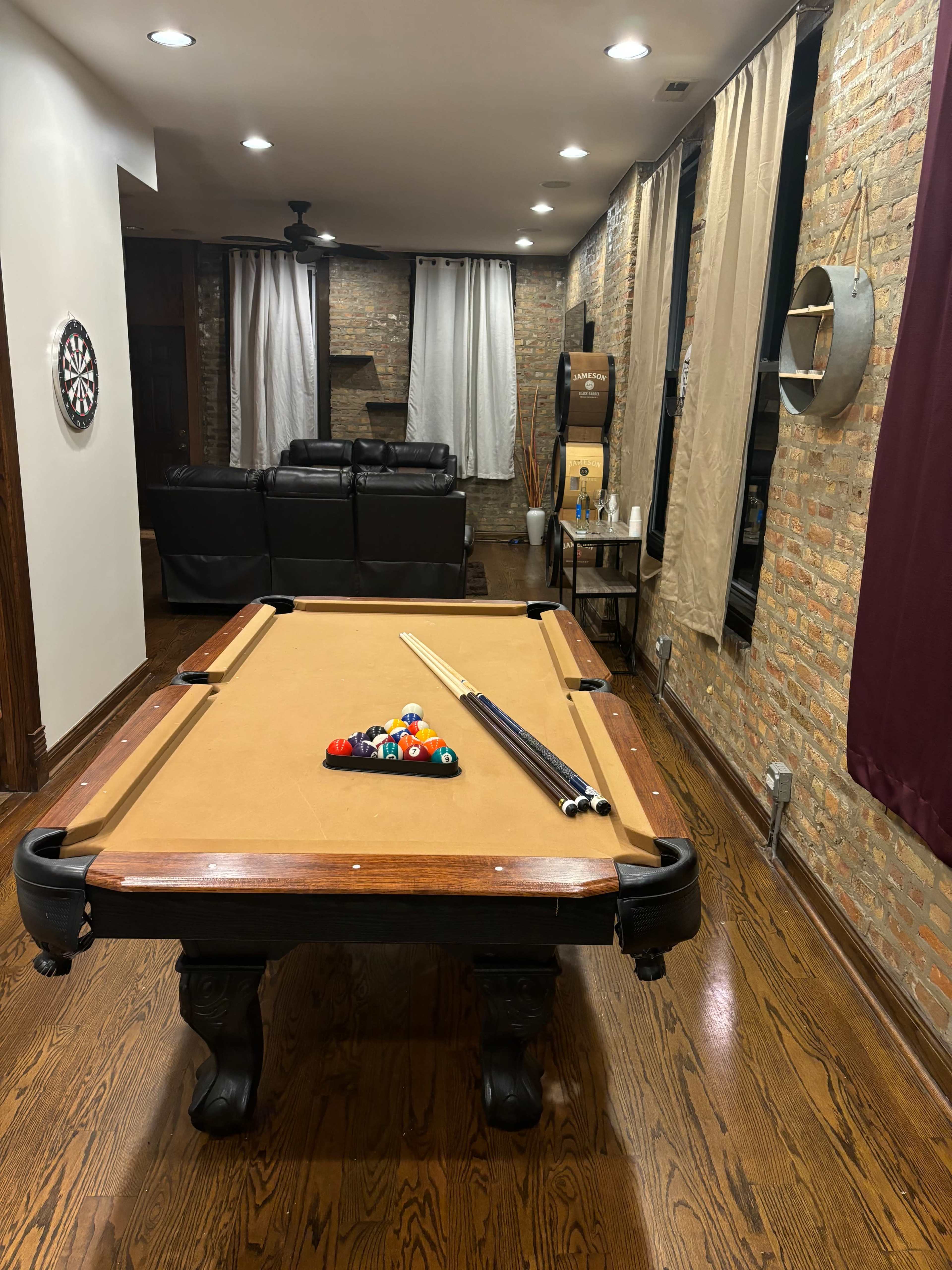 The image shows a billiards table in a room with wooden flooring, gray walls, and various furnishings, including a dartboard and a couch.