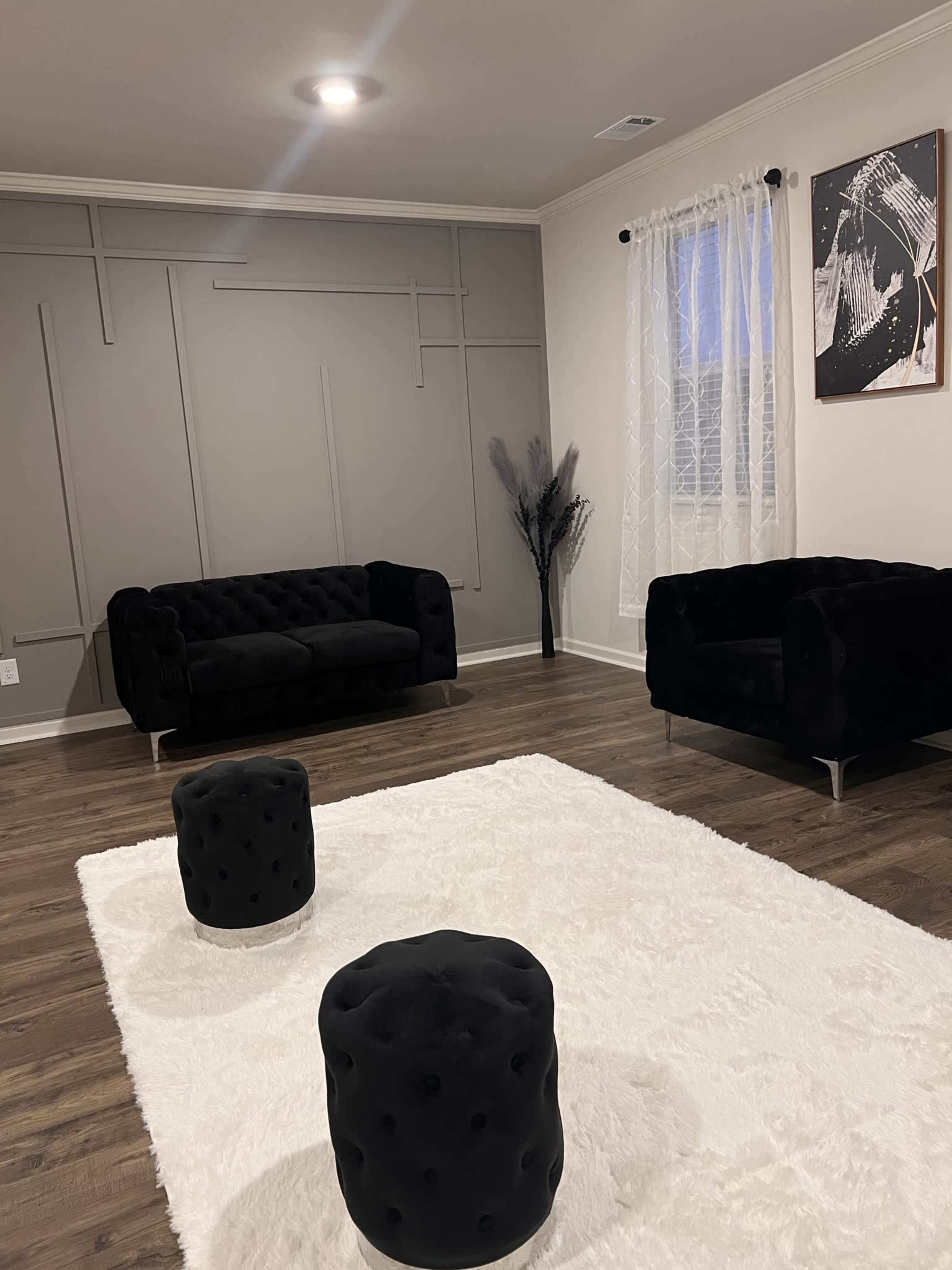A living room with a gray wall, a white area rug, a black tufted sofa, a matching armchair, and decorative black stools.
