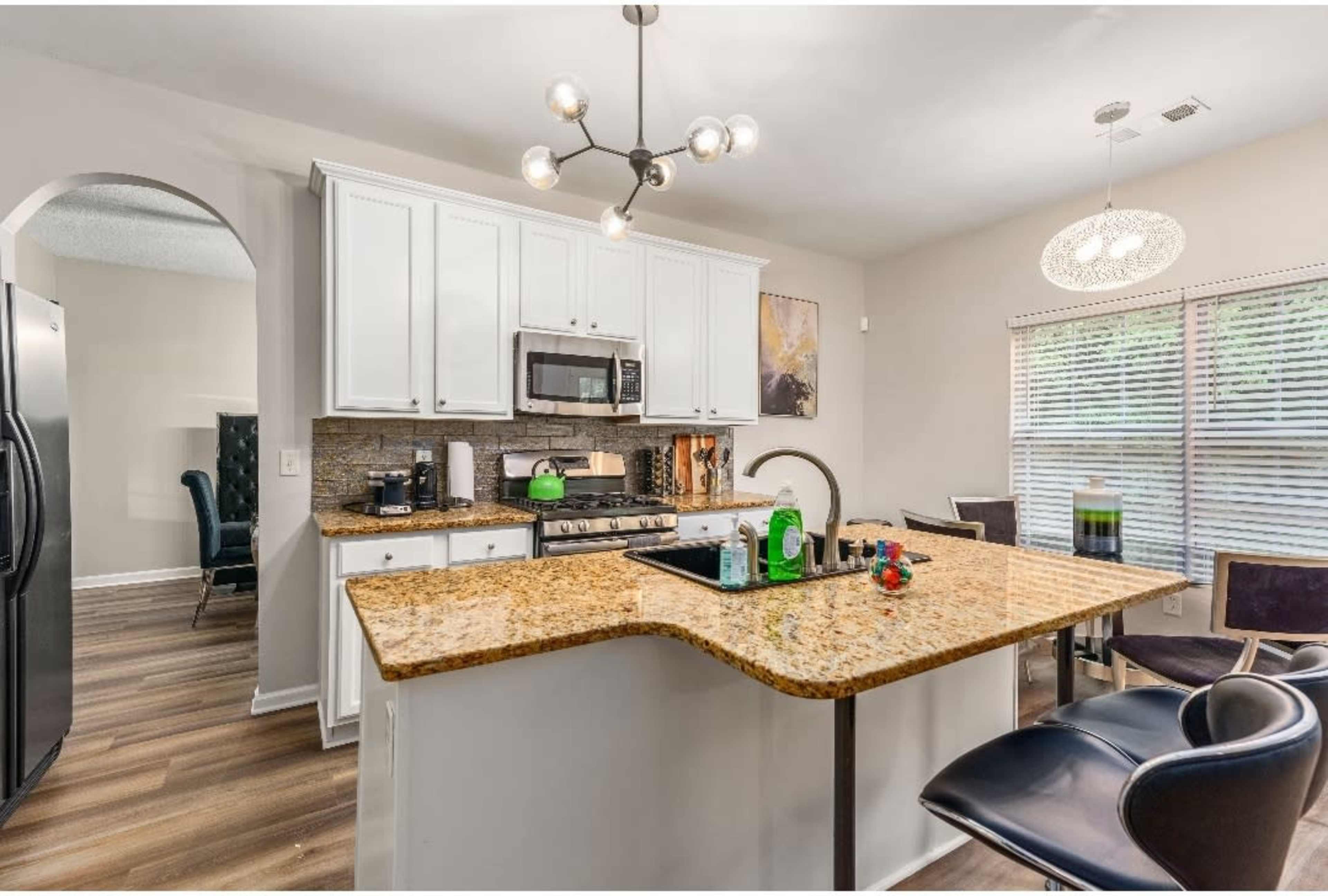 A modern kitchen with white cabinets, a granite countertop, stainless steel appliances, and a dining area featuring a round light fixture.