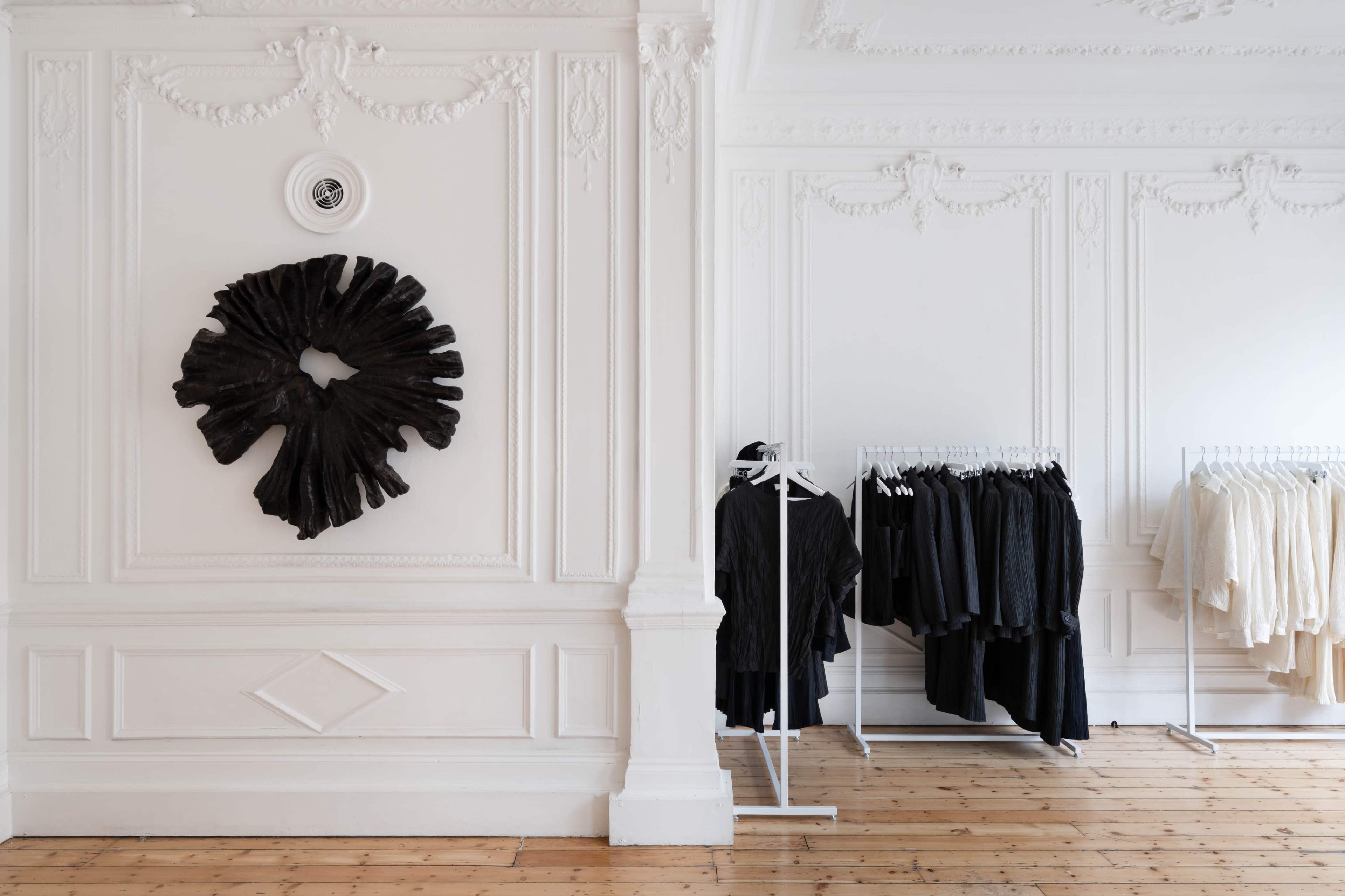 The image shows a minimalist clothing display in a white-walled room featuring a dark artistic wall piece and racks of clothing in neutral colors.