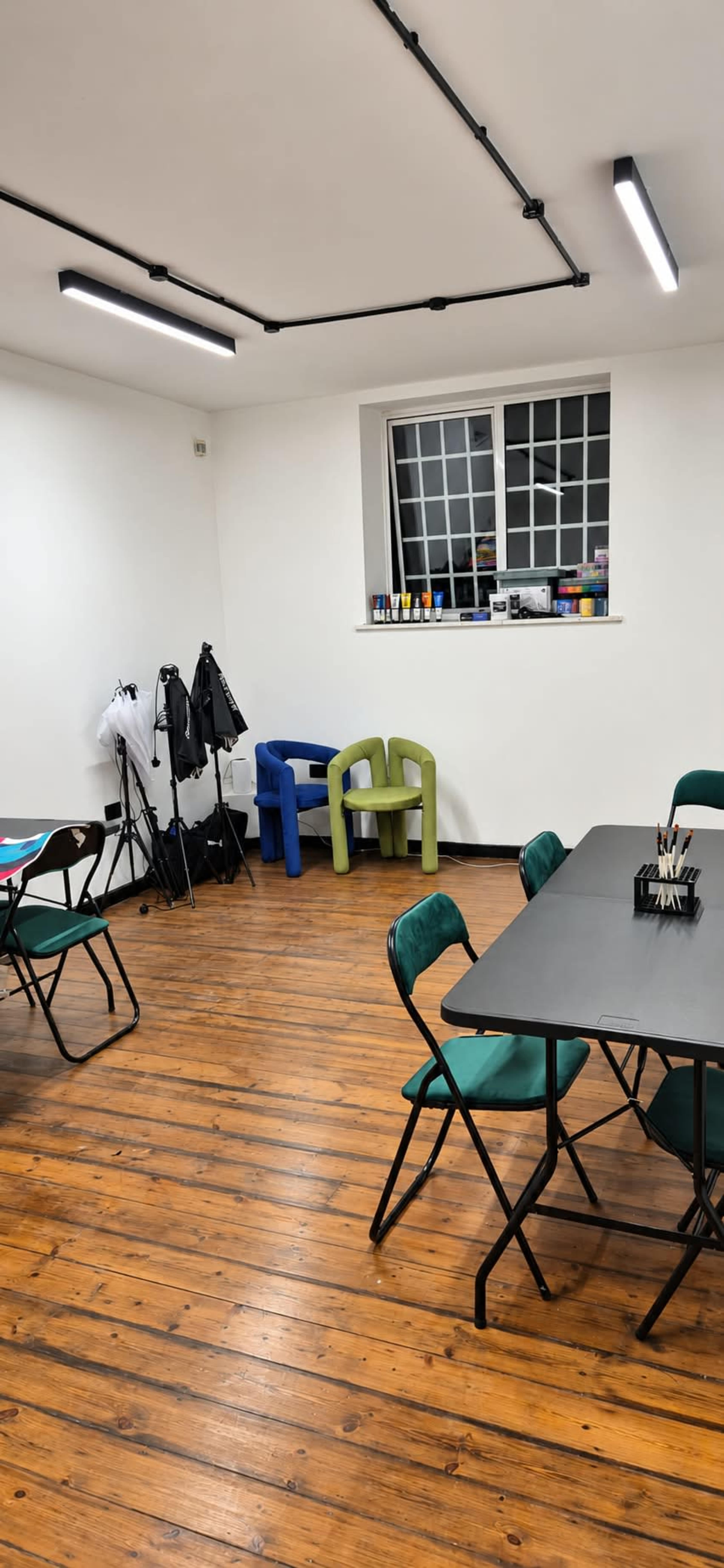The image shows a brightly lit room with wooden floors, featuring several folding chairs and tables, along with a small green and blue chair set against a wall equipped with shelves.
