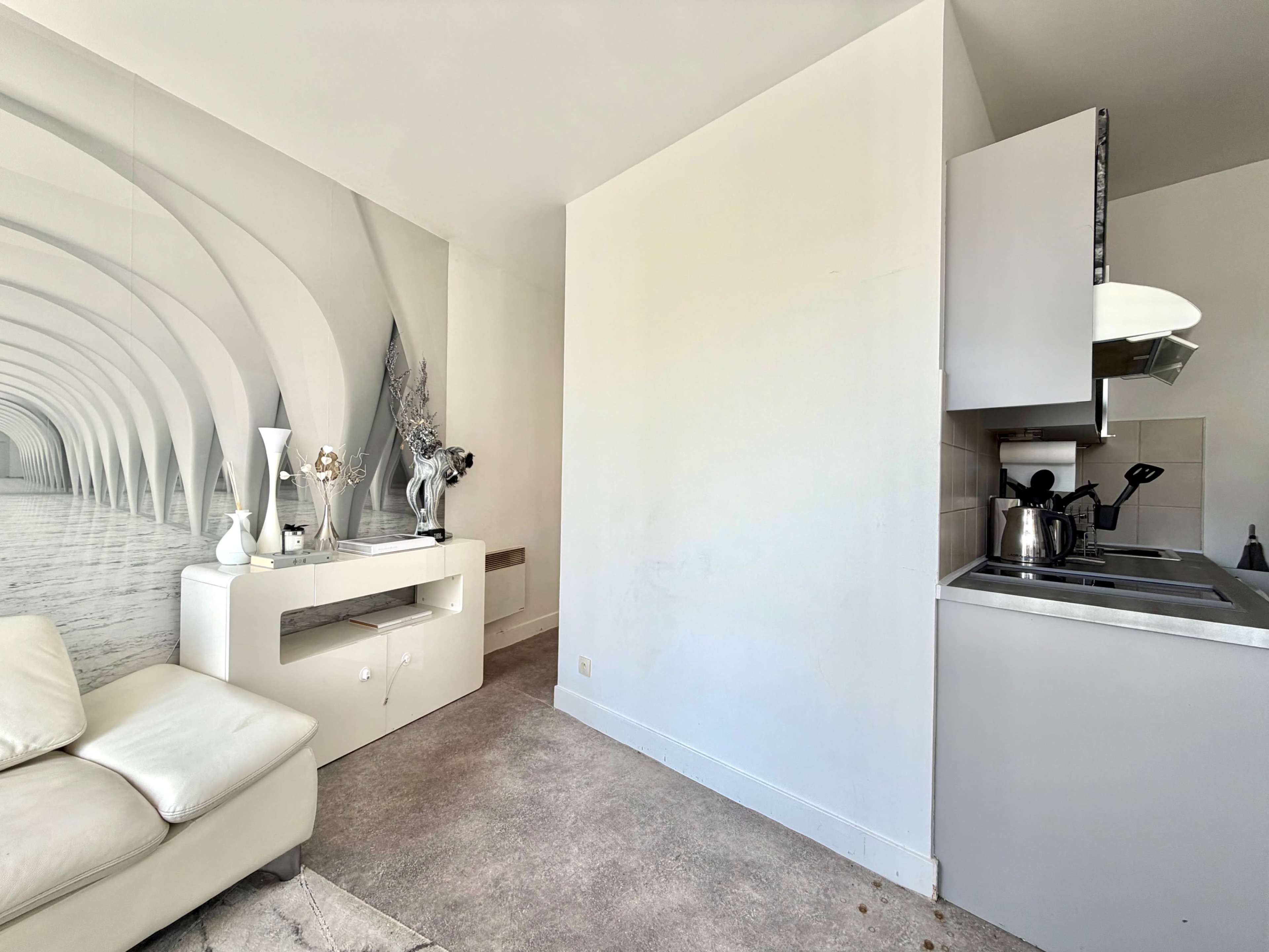 A modern, minimalist living space features a white couch, a decorative table, and an adjoining kitchenette with a stove and sink.