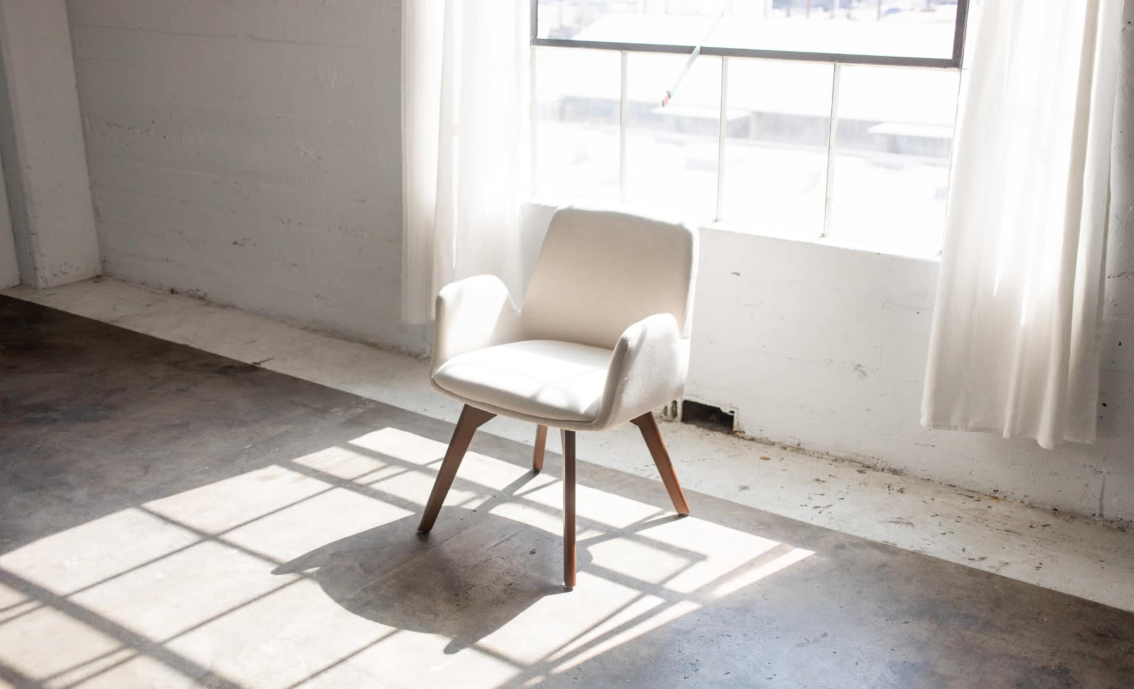A light-colored chair with wooden legs is positioned in front of a large window with white curtains, illuminating the surrounding concrete floor.