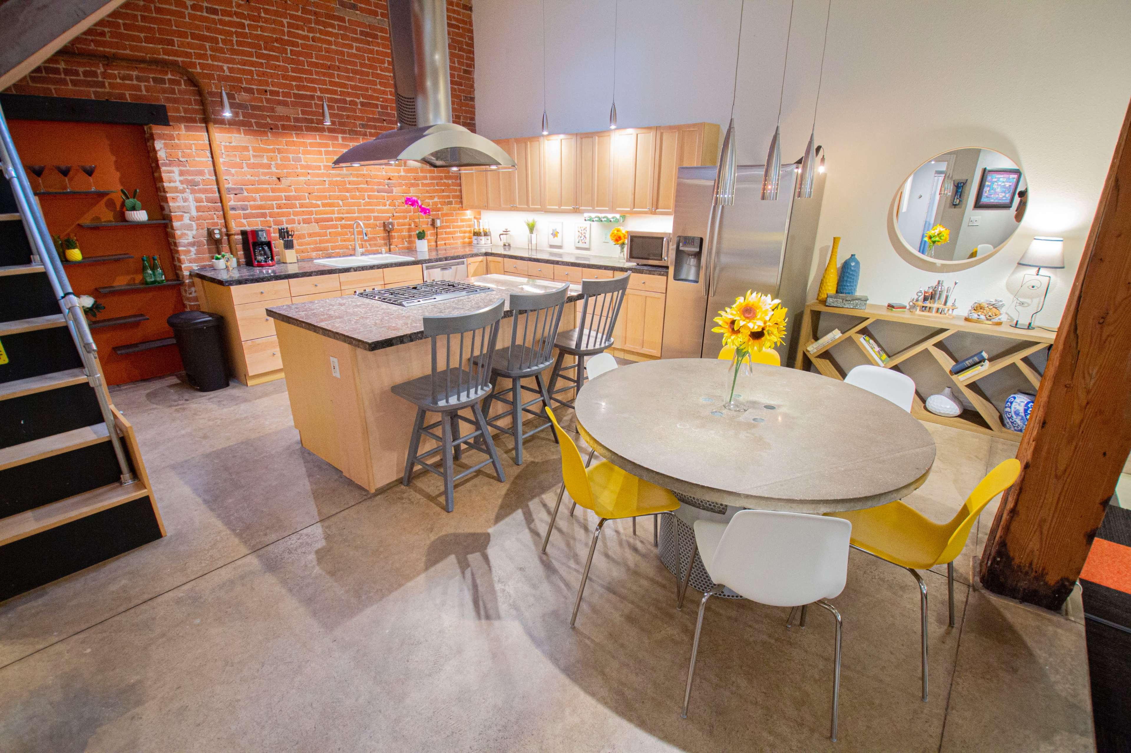 A modern kitchen and dining area with a large circular table, yellow chairs, and a brick wall backdrop.