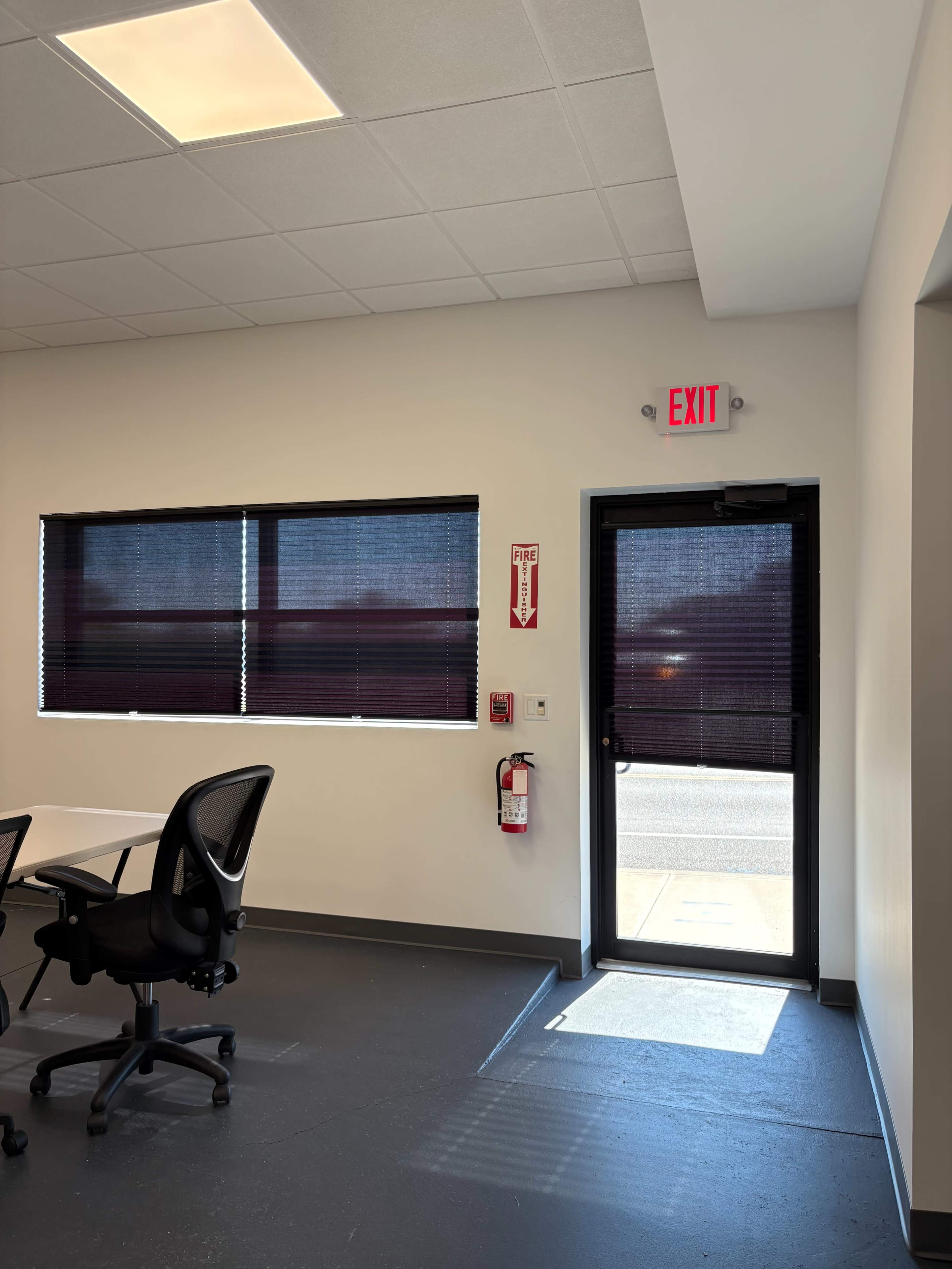 The image shows the interior of a room with a door leading outside, along with a window covered by blinds, a fire extinguisher, and office chairs set around tables.