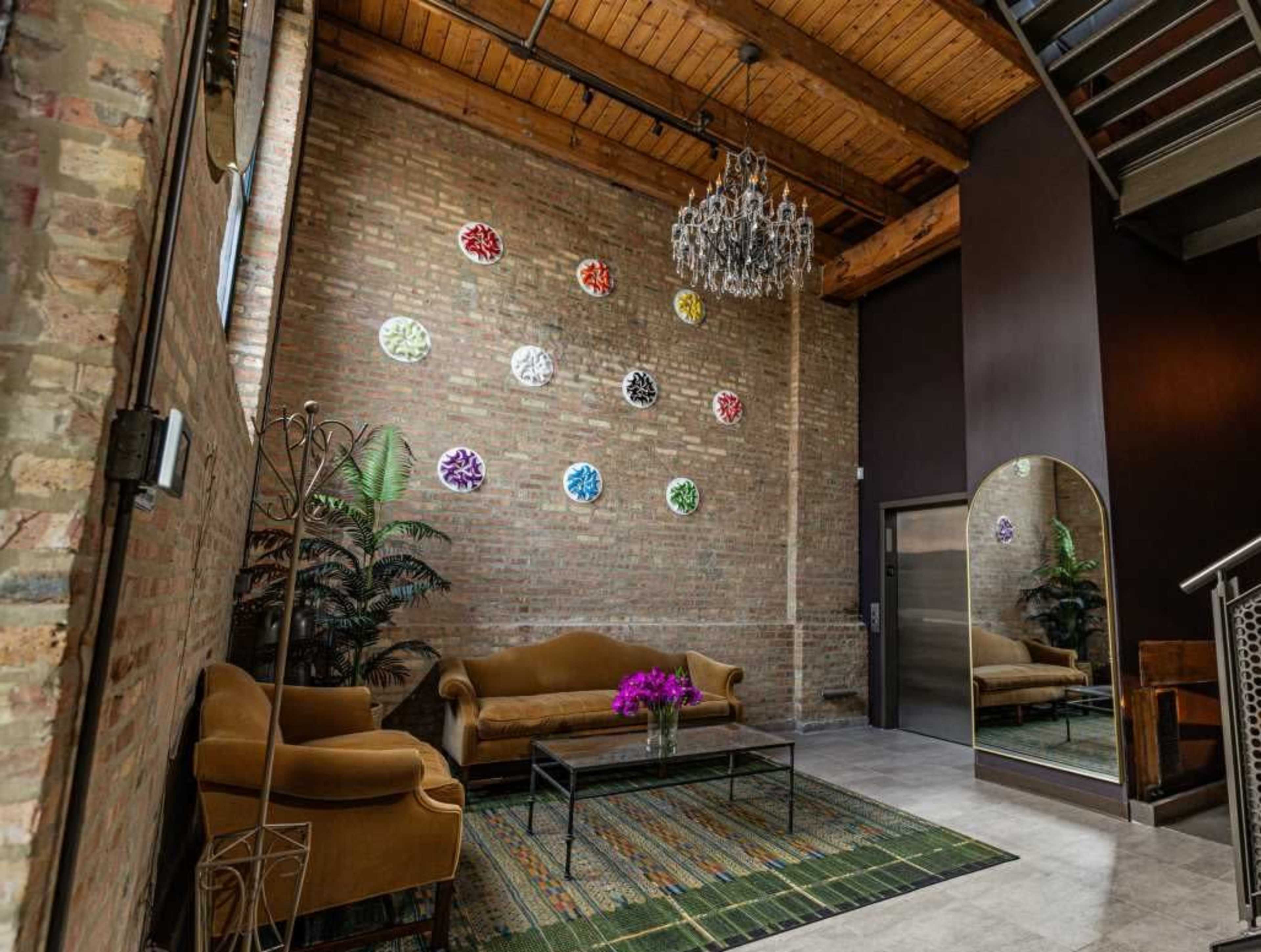 A cozy entryway features a brown sofa, a coffee table with flowers, a mirror, and colorful wall decor against a brick wall.