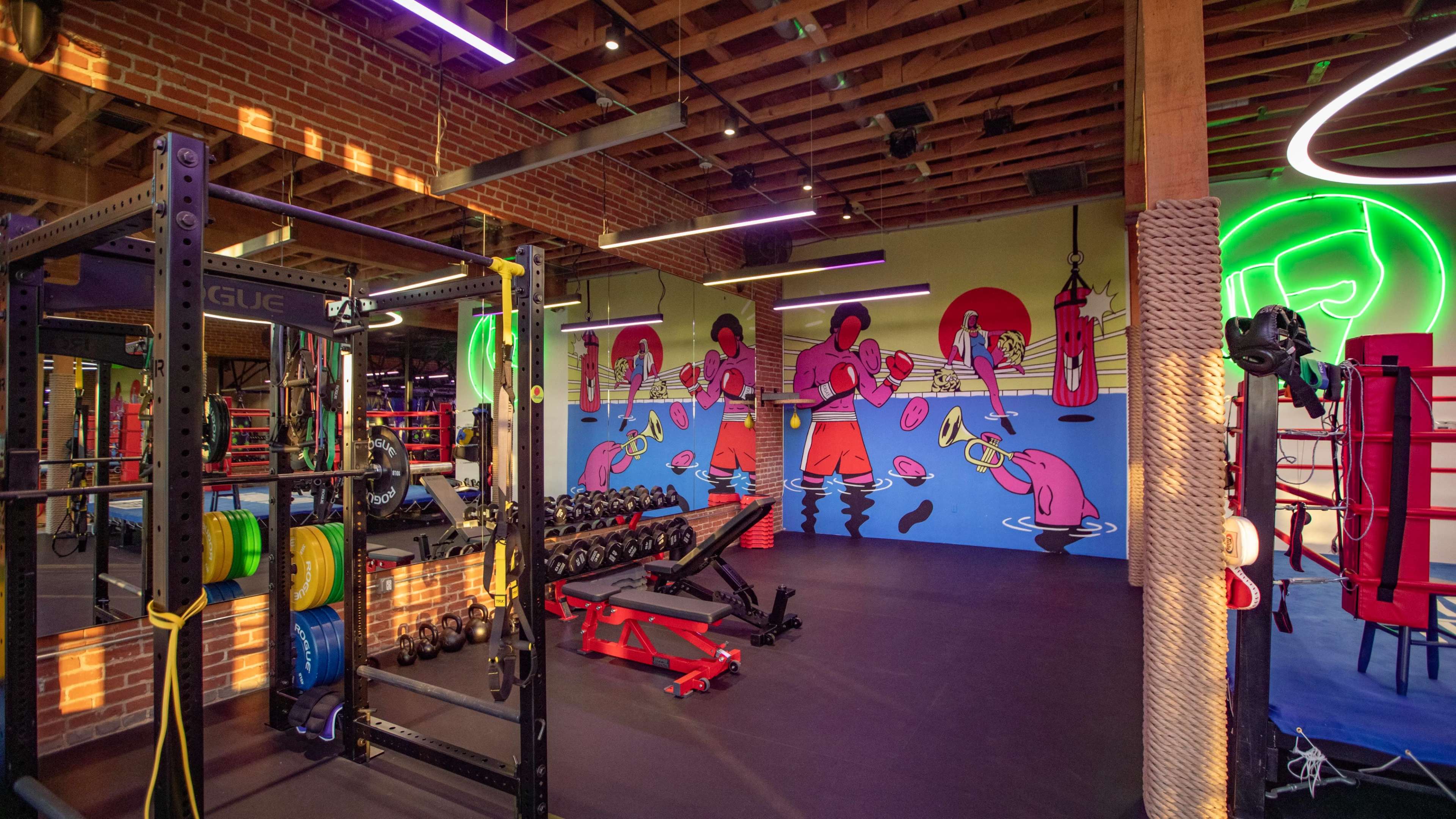 A modern gym interior featuring weightlifting equipment, a colorful mural of boxers on the wall, and various boxing gear.