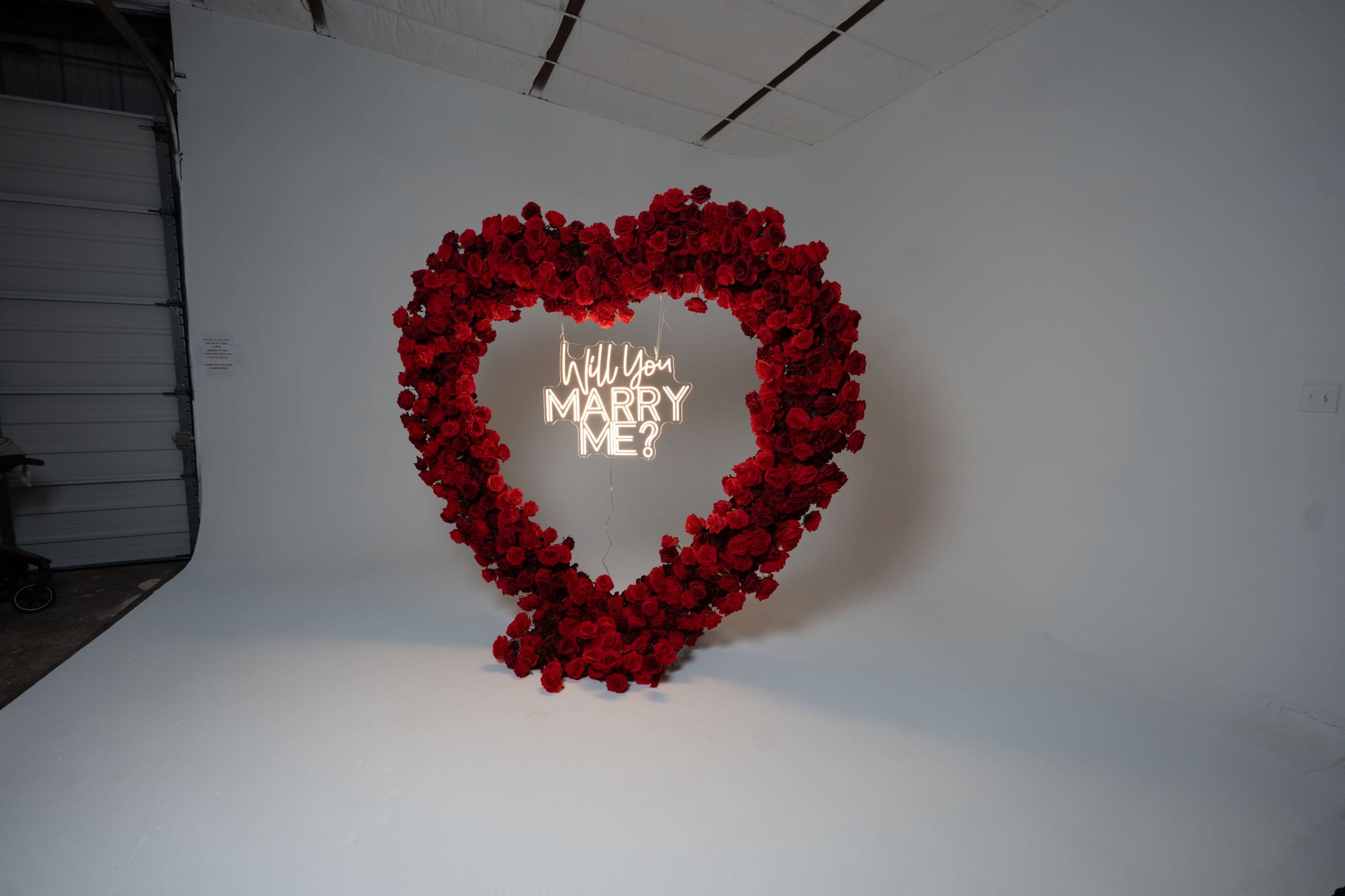 A large heart-shaped arrangement of red roses surrounds a lit sign that reads, "Will You Marry Me?" in a simple white font against a plain background.