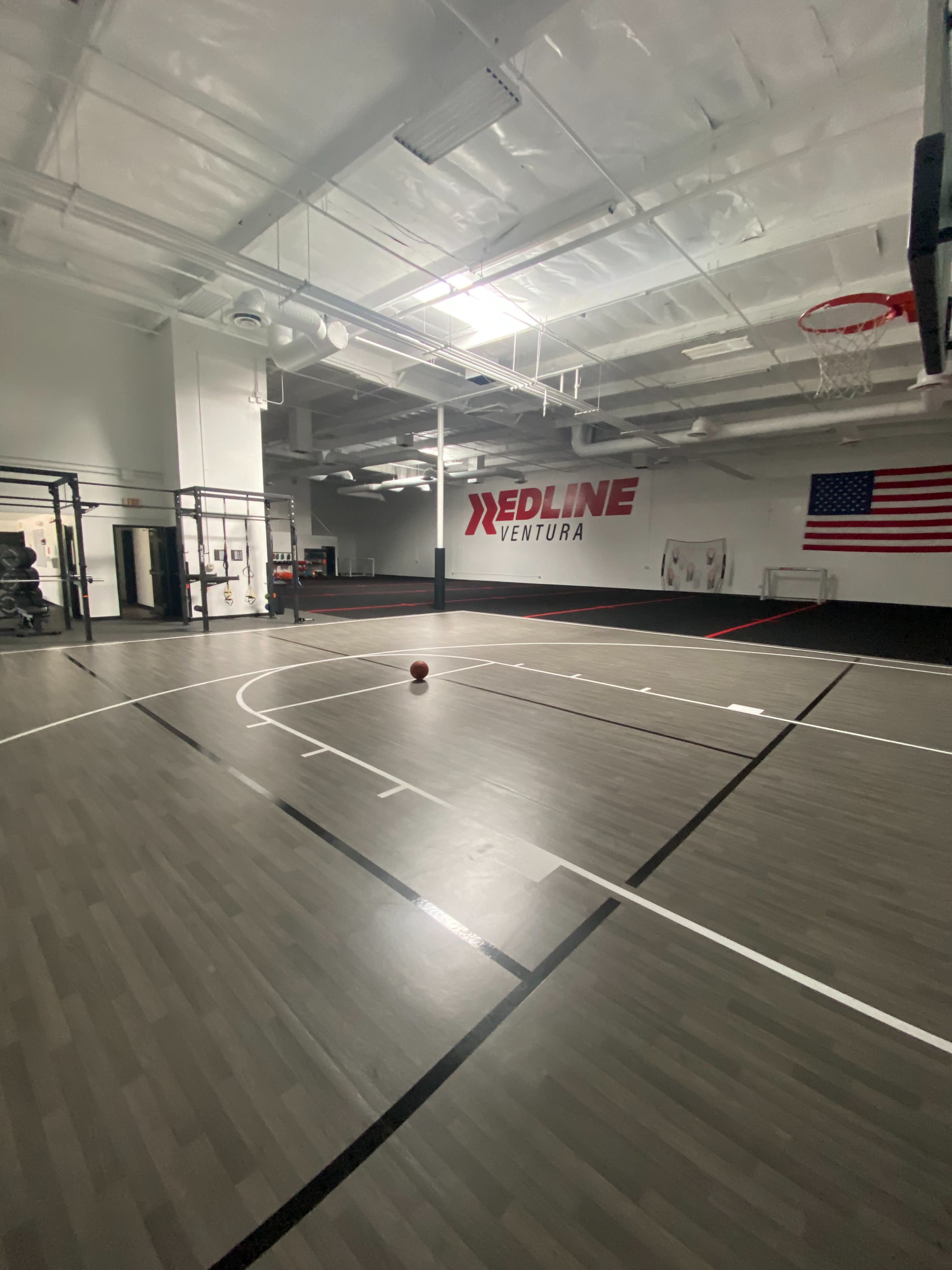 The image shows an empty gymnasium with a basketball on the court and a large American flag on the wall.
