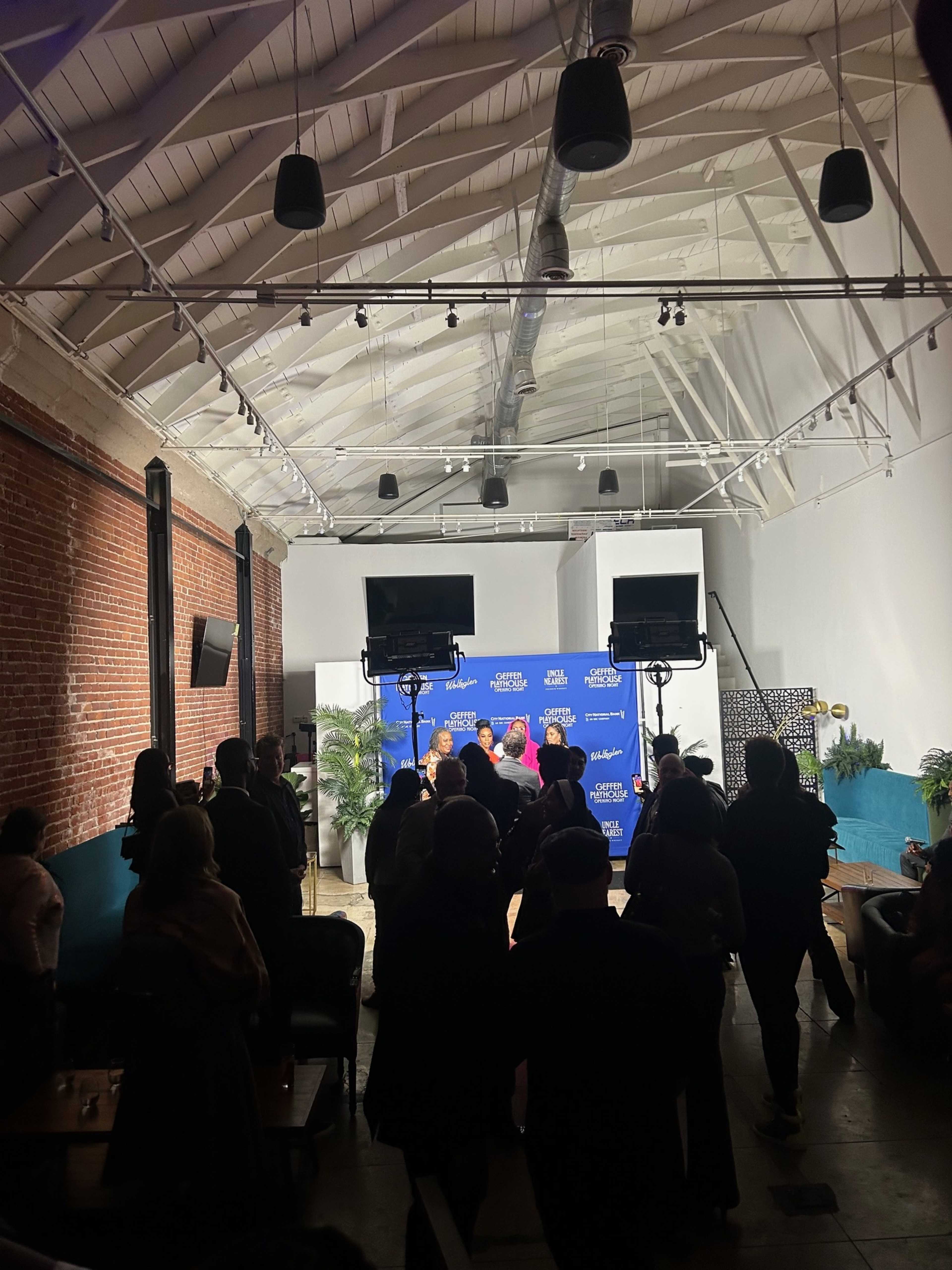 A group of people is gathered in a well-lit venue with a blue backdrop, while two cameras are positioned in the foreground.
