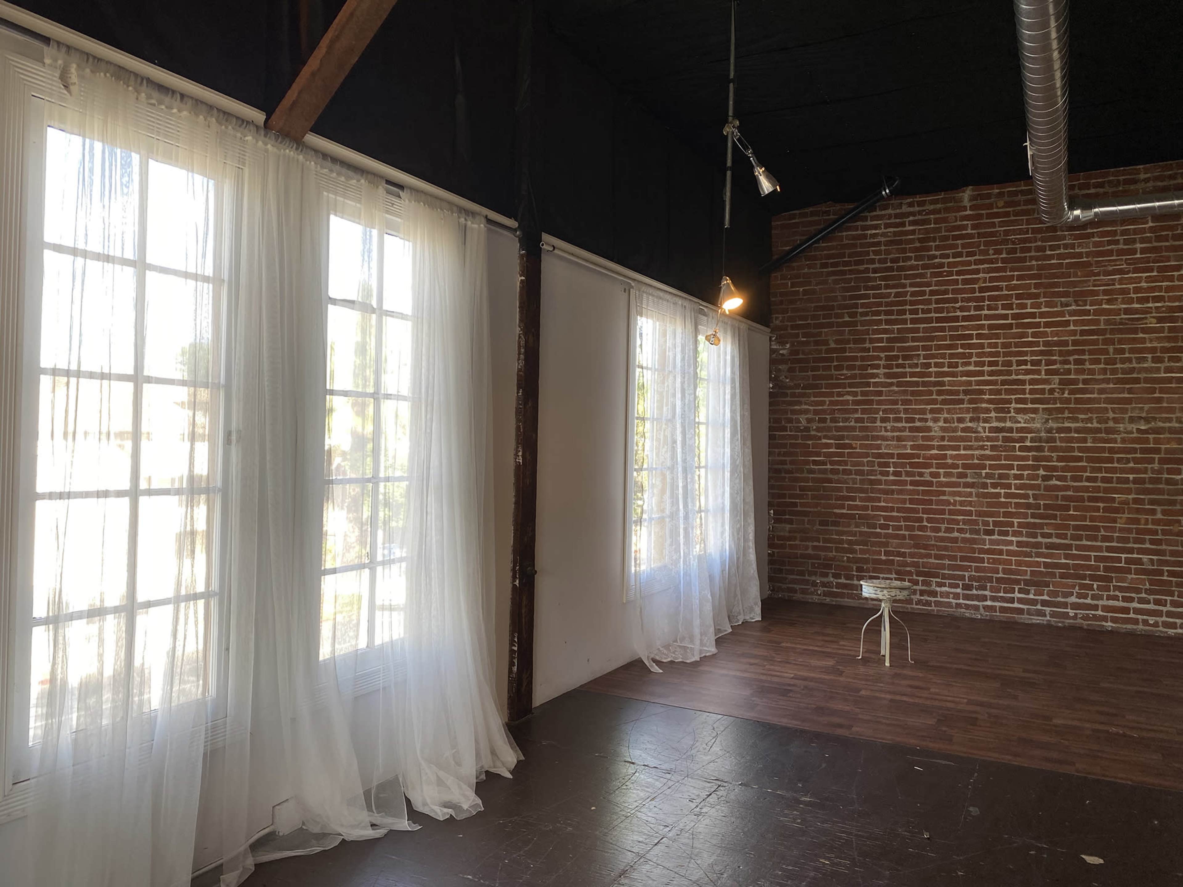 The image shows an interior space with large windows draped in sheer curtains, exposing brick walls and a wooden floor.