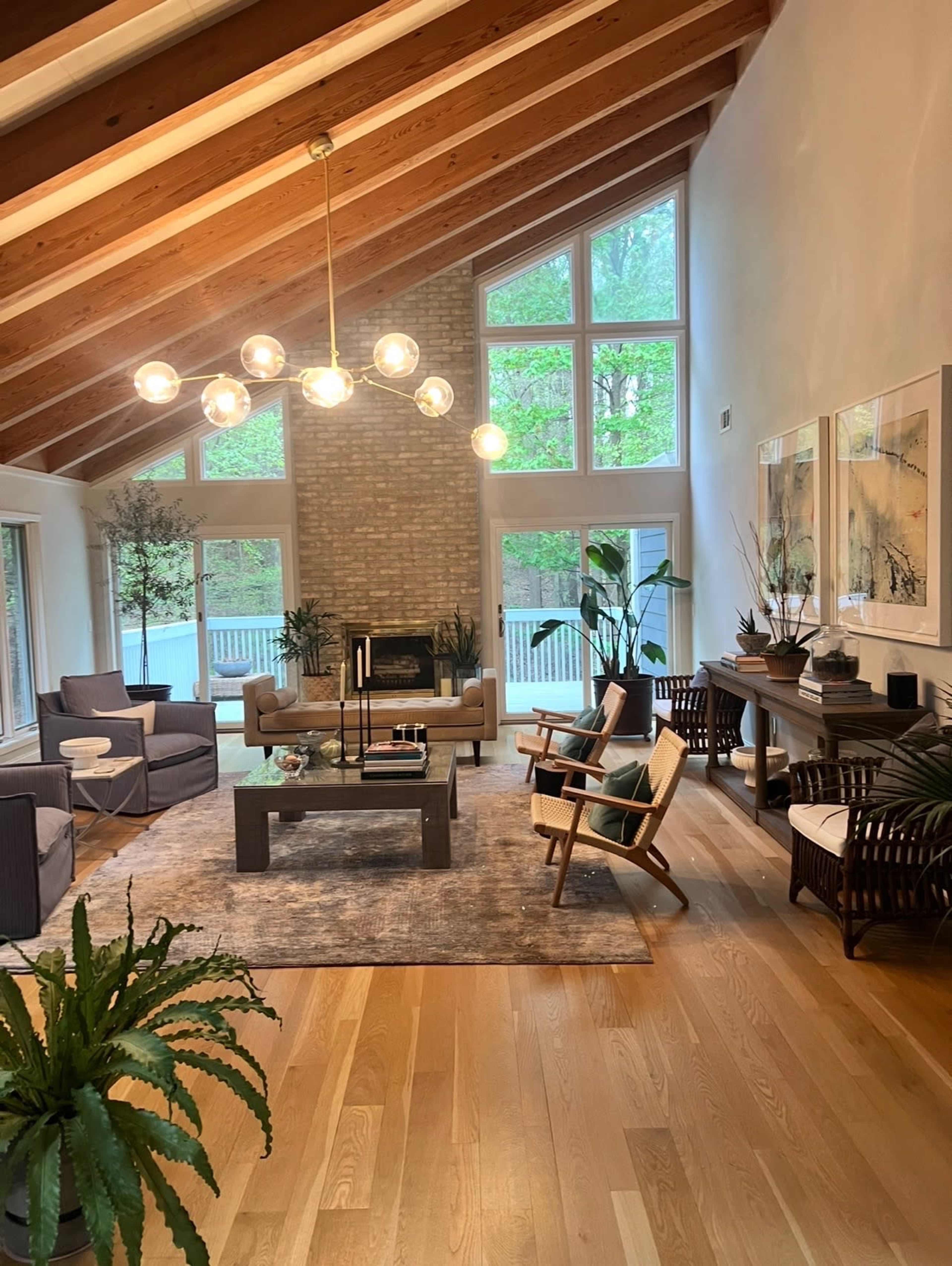 The living room features a light wood floor, a high-beamed ceiling, large windows, and modern furniture arranged around a coffee table.